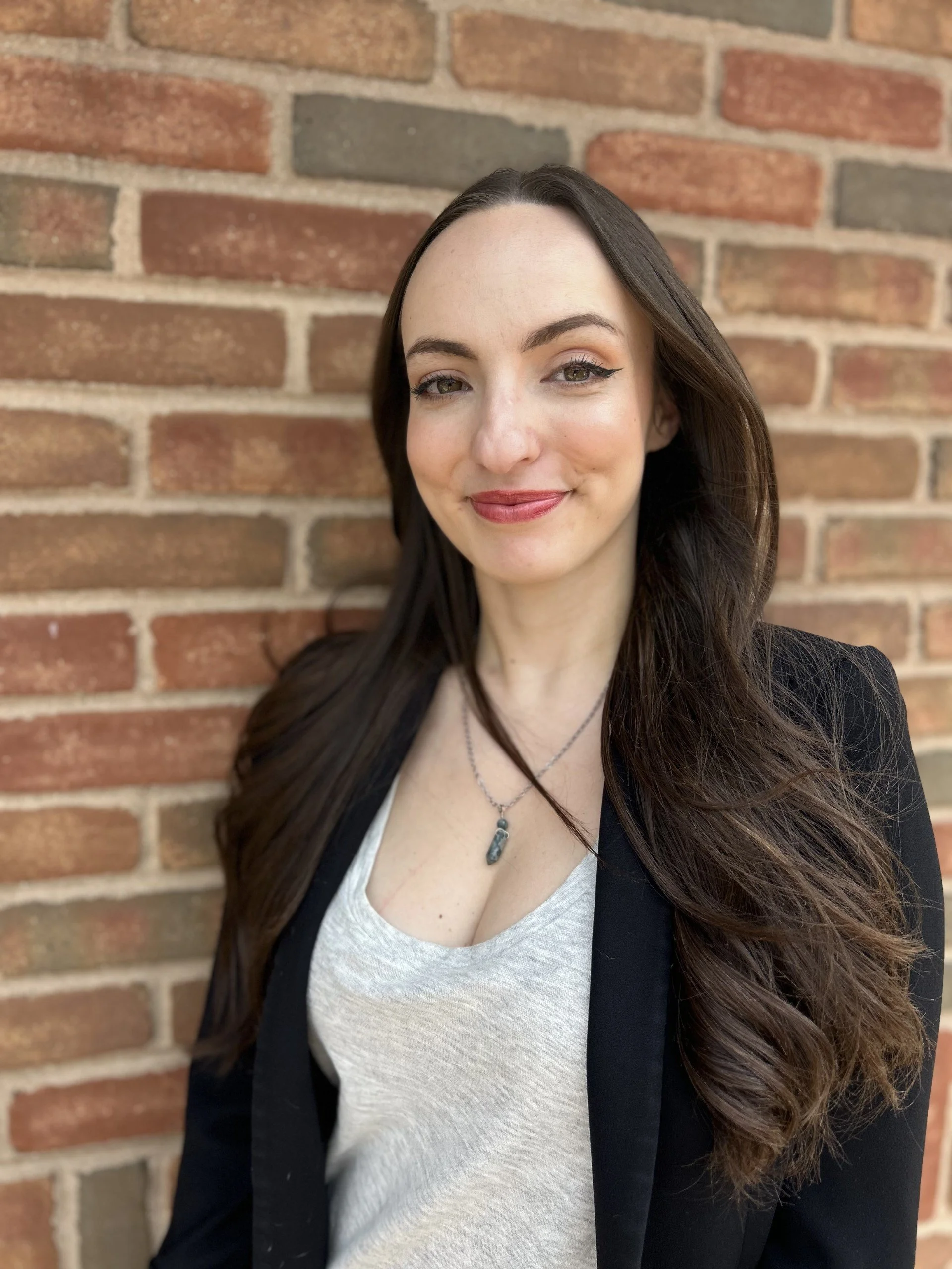 A woman with long dark hair, wearing a black blazer over a light gray top, standing against a brick wall, smiling at the camera.