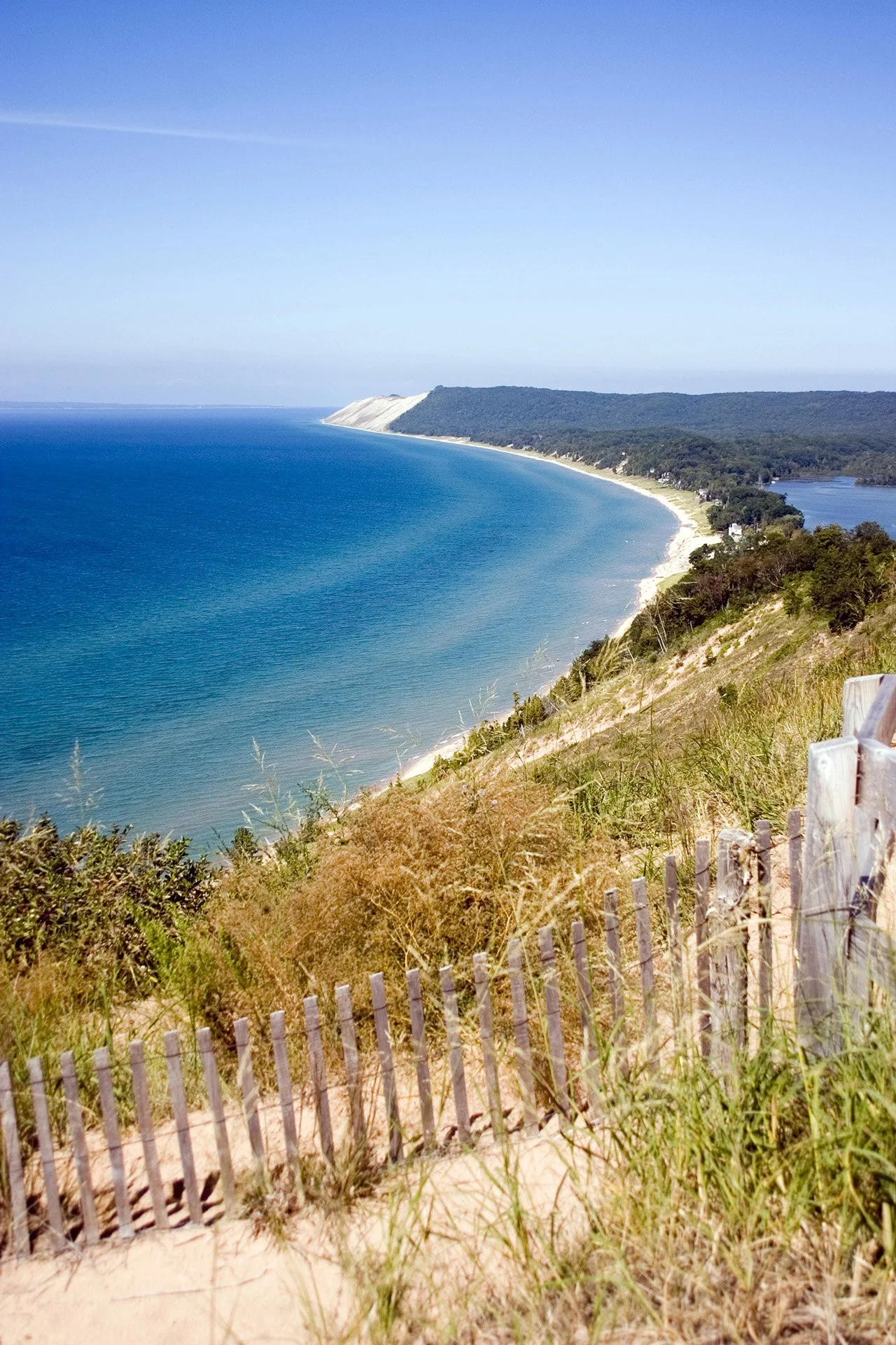 A coastal landscape with a sandy beach, clear blue ocean, green forested hills, and a bright sky.
