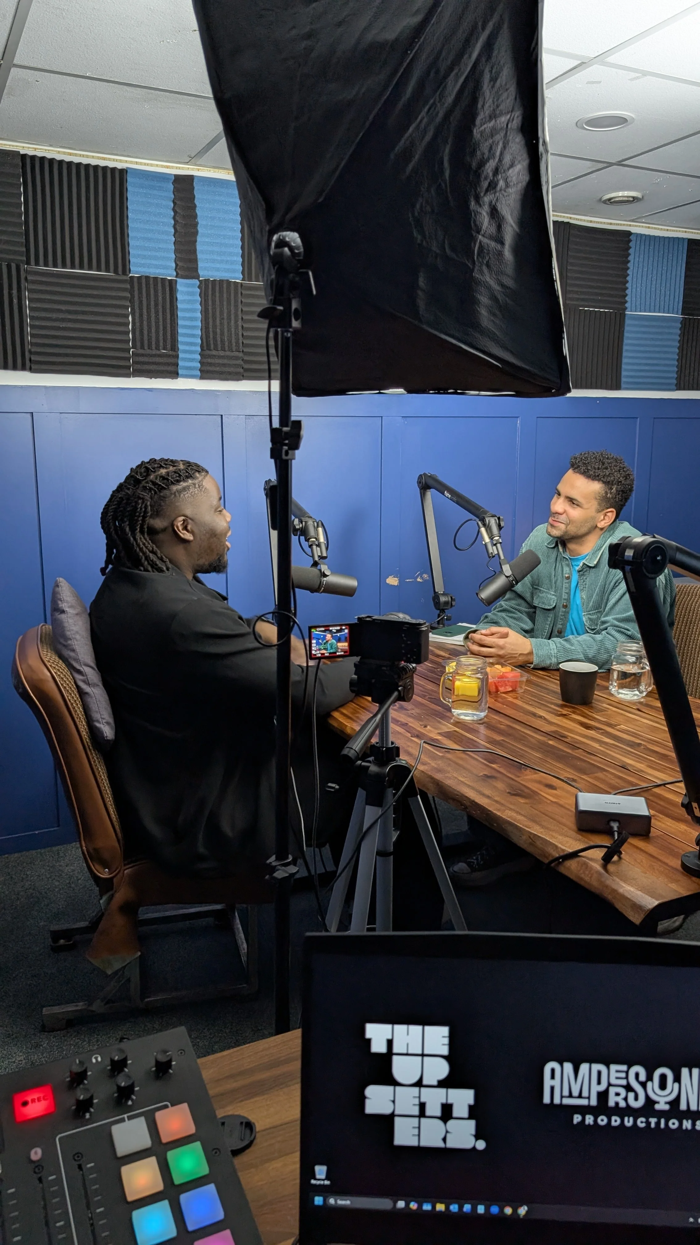 Two men are having a conversation in a podcast recording studio with microphones, a camera, and recording equipment on the table. One man is wearing a black jacket, and the other is wearing a green jacket. There are drinks on the table. A monitor screen displays logos.
