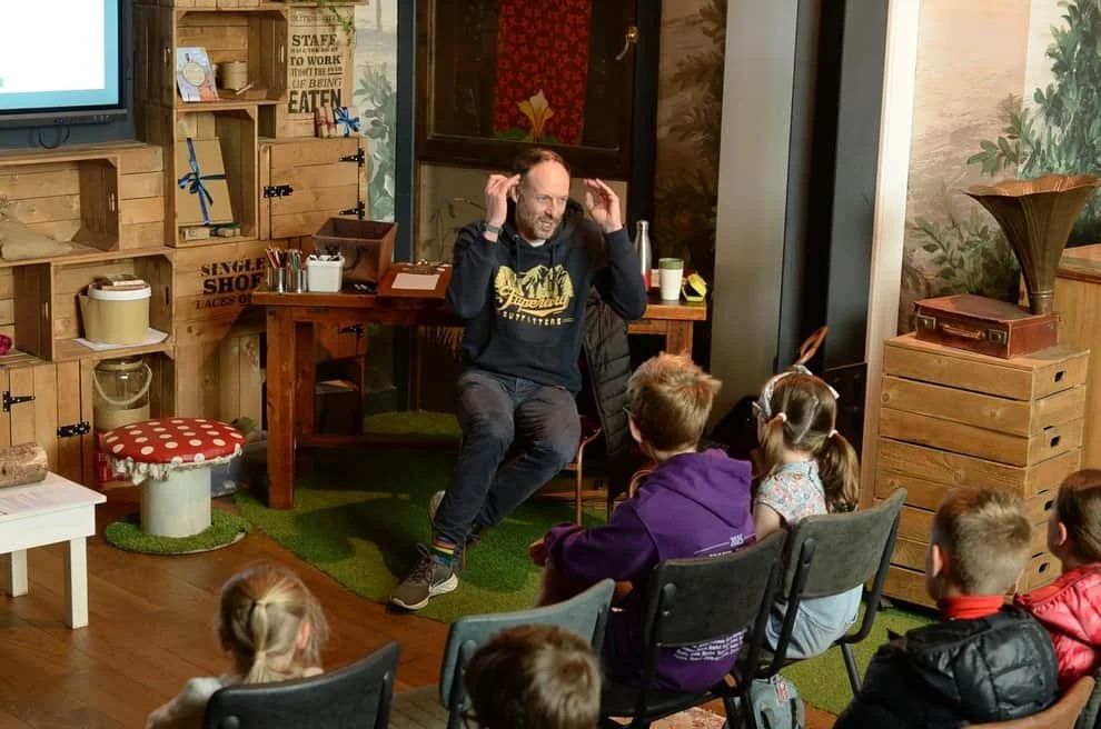 A workshop for children led by writer TomJordan, part of Grimm & Co's Festival of Stories, in a cozy, rustic room with wooden shelves, a leopard print wallpapered door, and various decorative items.