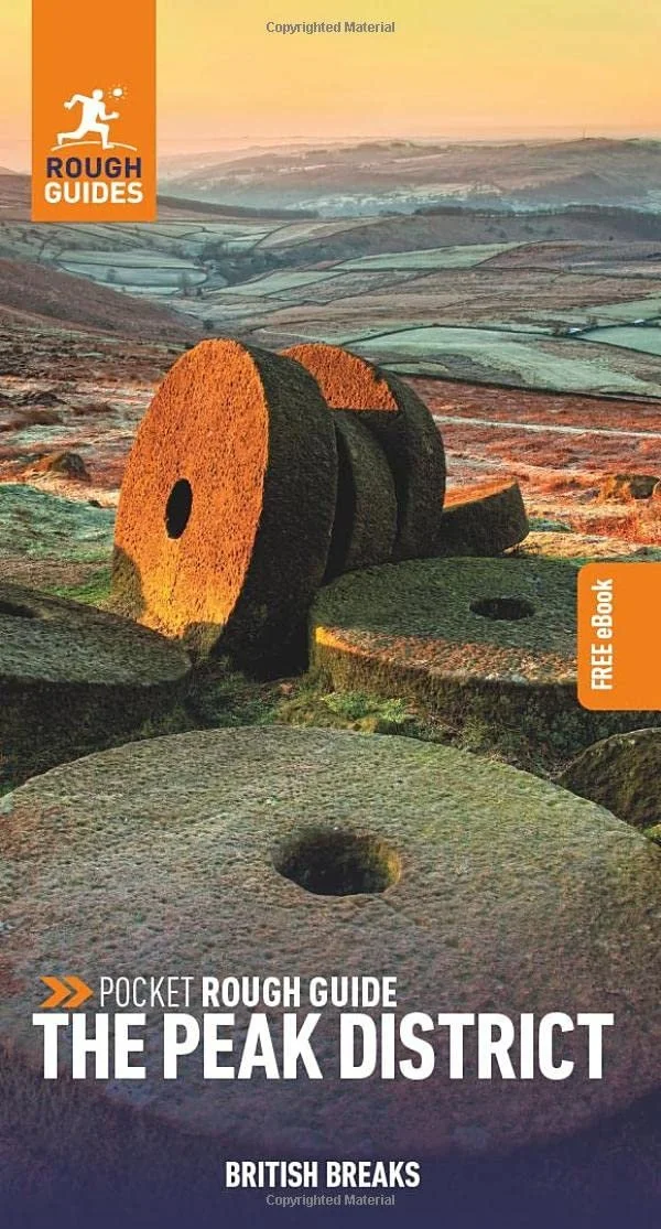 The cover of the travel guidebook 'Pocket Rough Guide: The Peak District', featuring large, ancient stone mill wheels on a hillside with rolling fields and hills in the background during sunset.