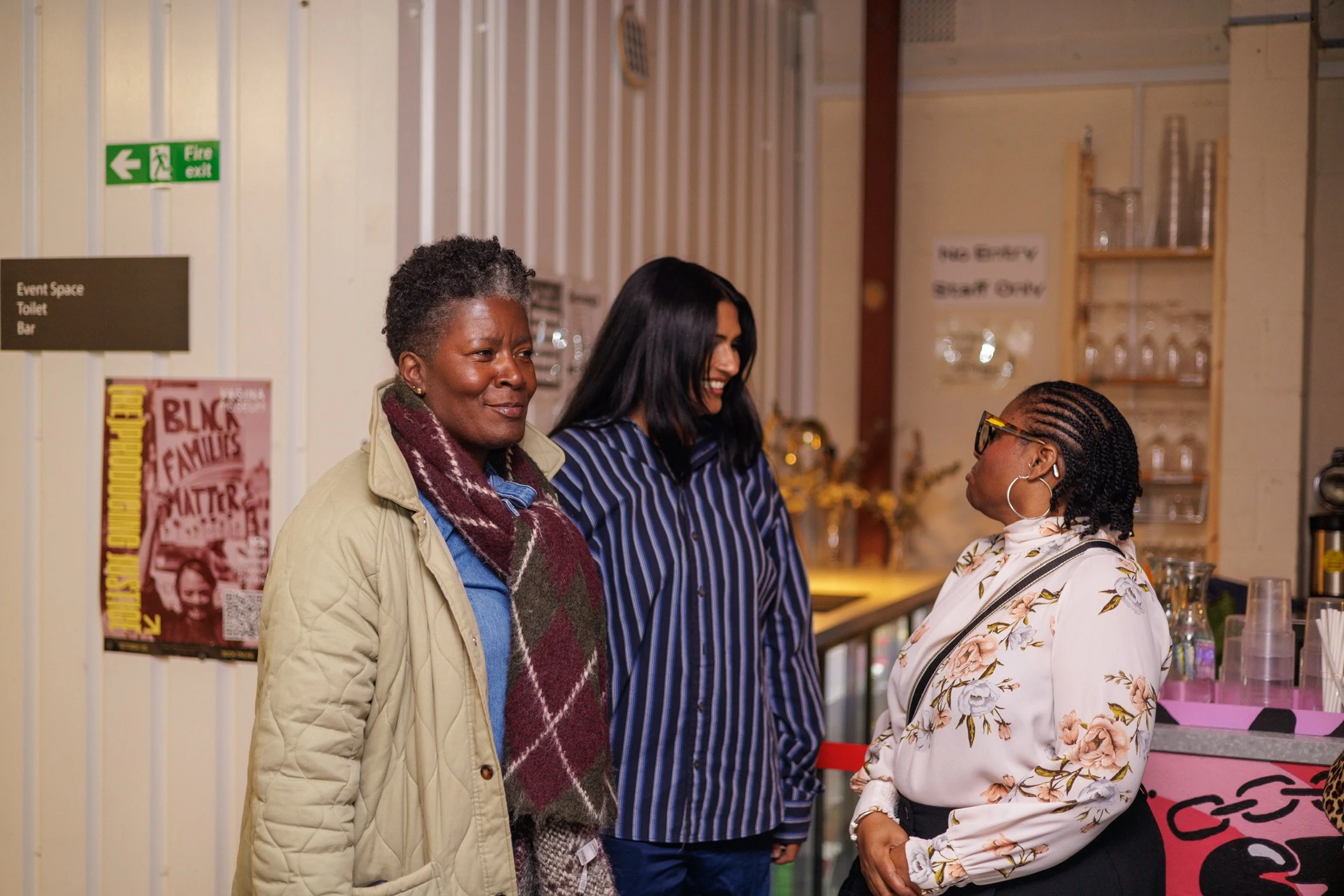 Three women engaged in conversation inside a room, with signs and shelves in the background.