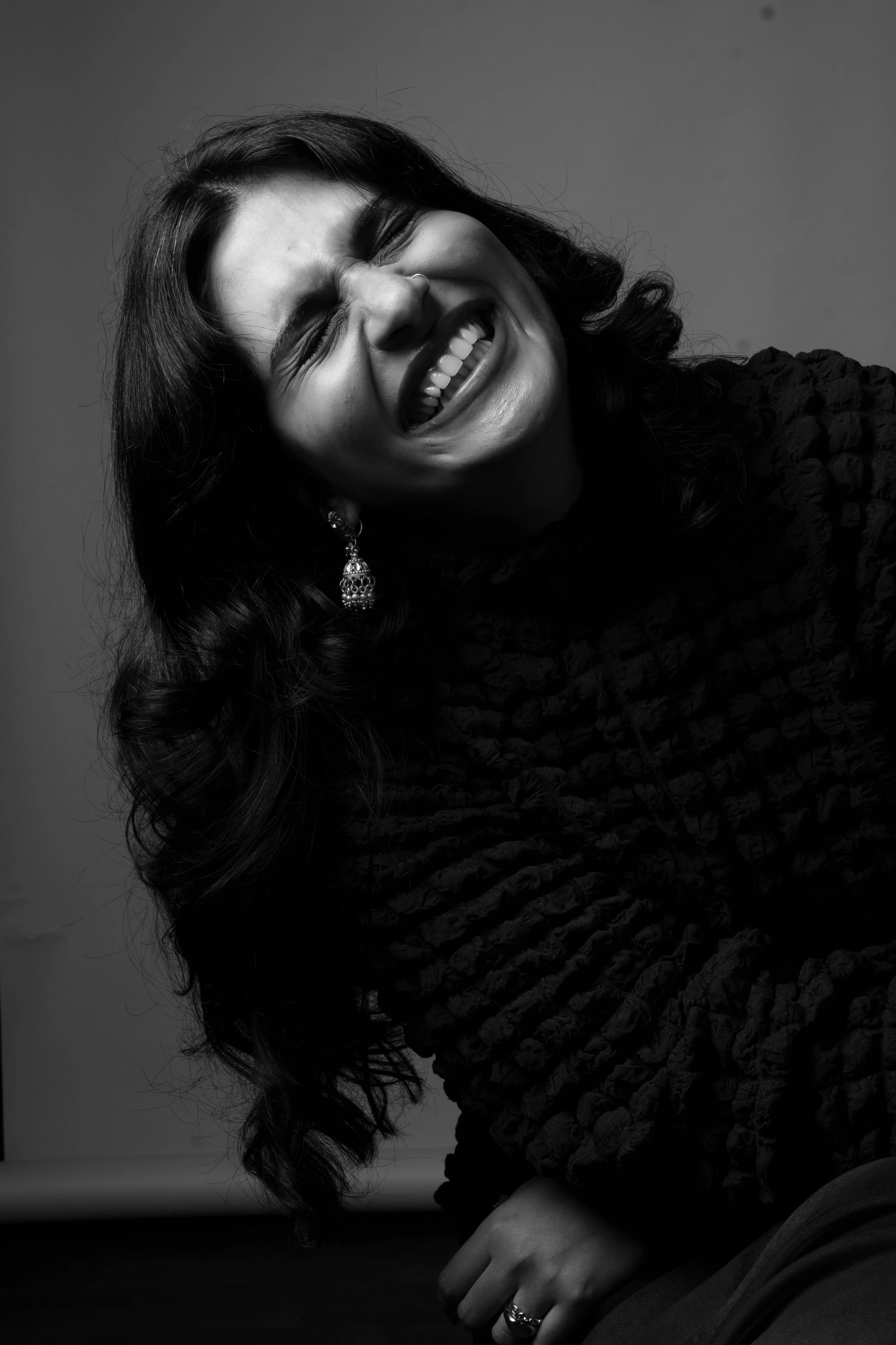 A woman with long dark hair, wearing earrings, is smiling and laughing in a black and white photo.