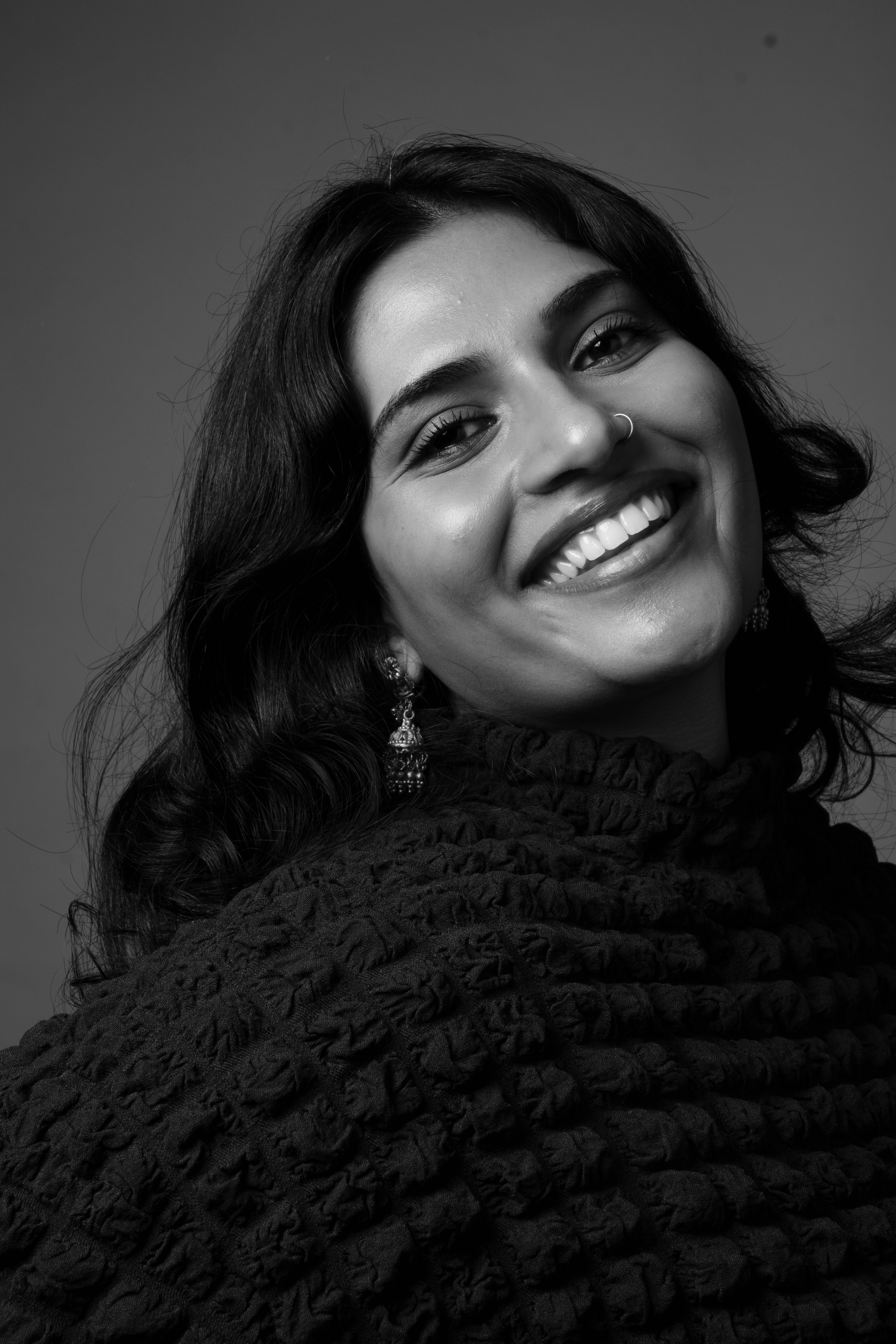 A smiling woman with dark wavy hair, wearing earrings and a textured garment, in black and white portrait photography.