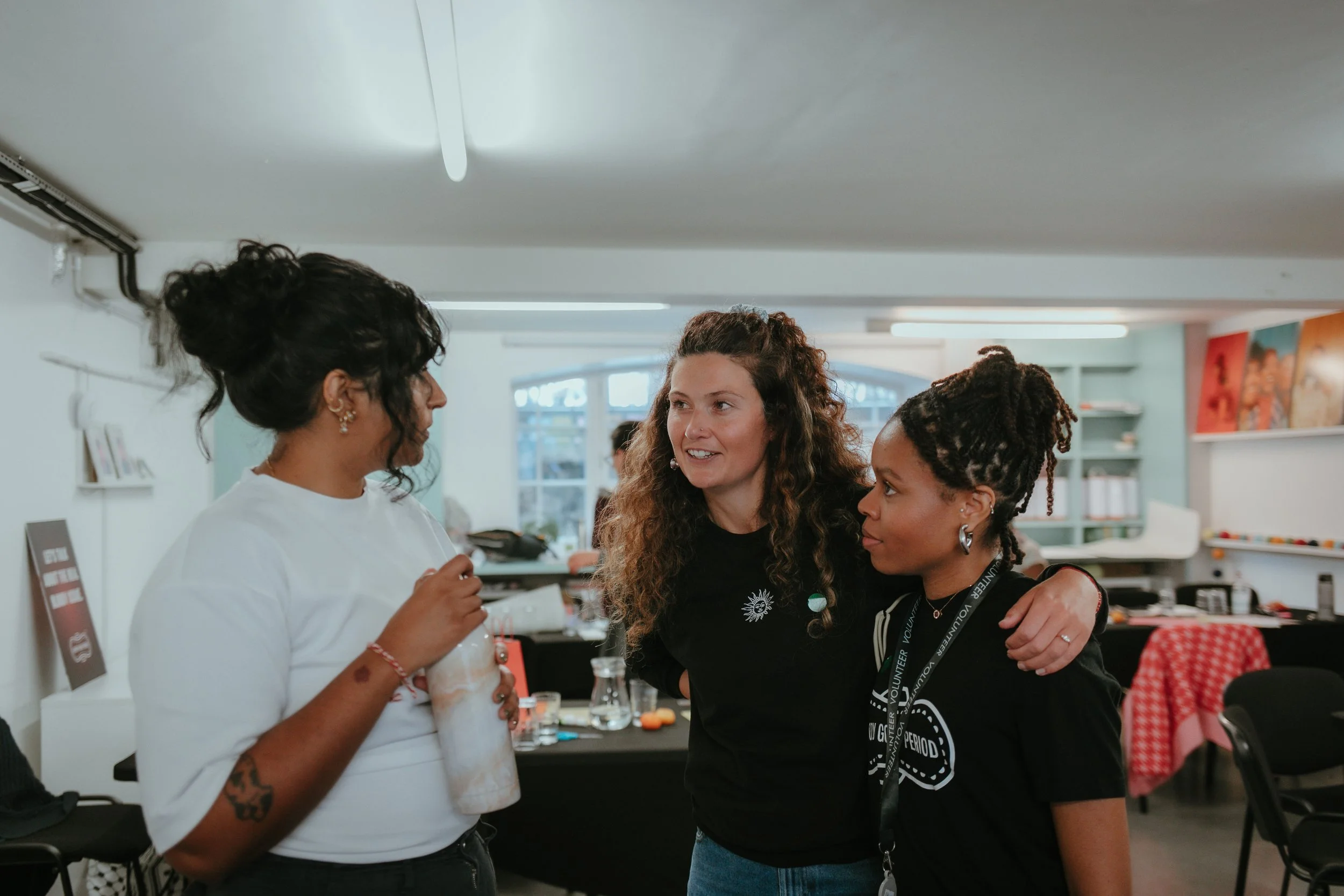 Three women talking in a bright room with shelves and colorful artwork in the background.