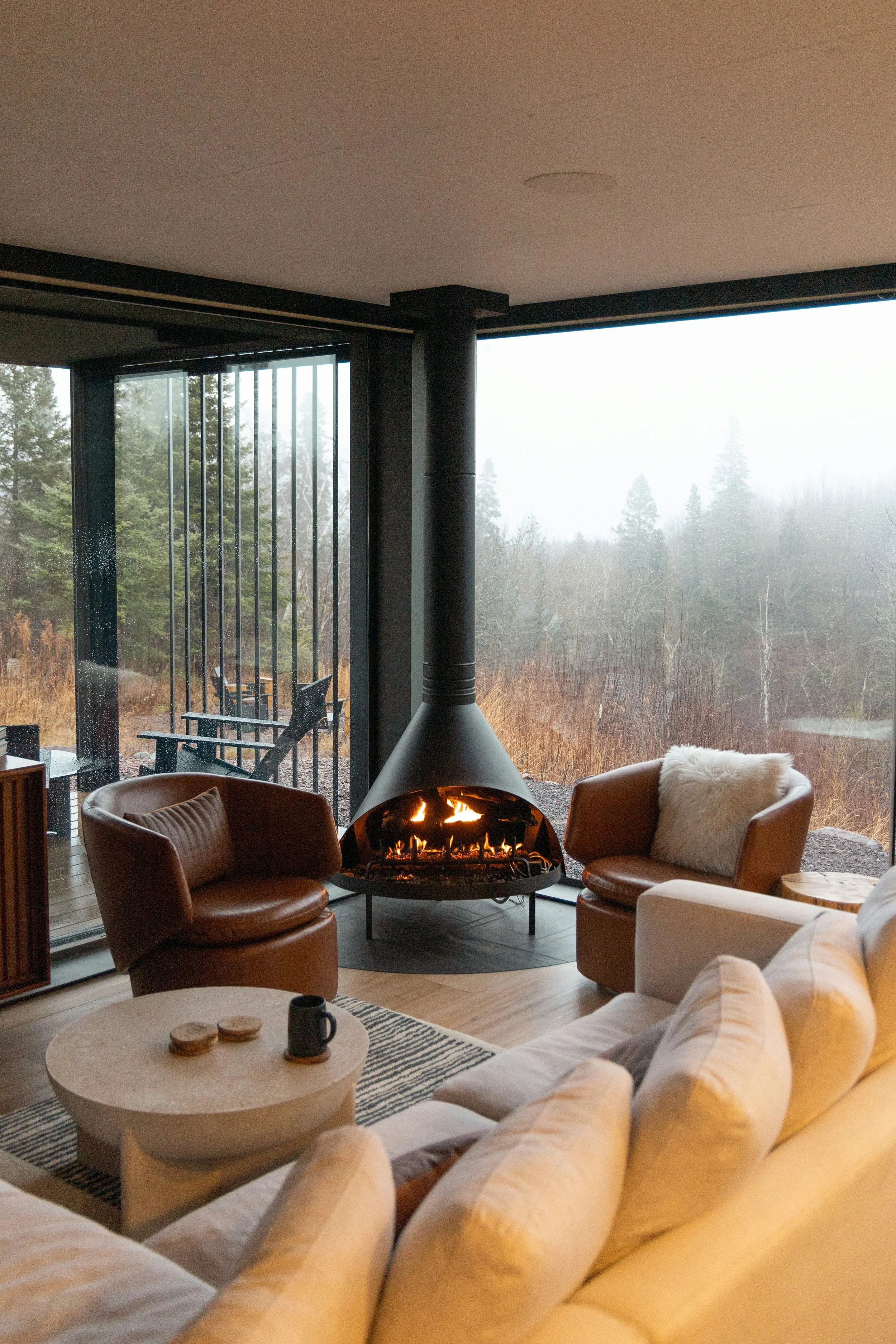 Living room with leather armchairs around a modern fireplace, view of foggy trees outside through large windows.