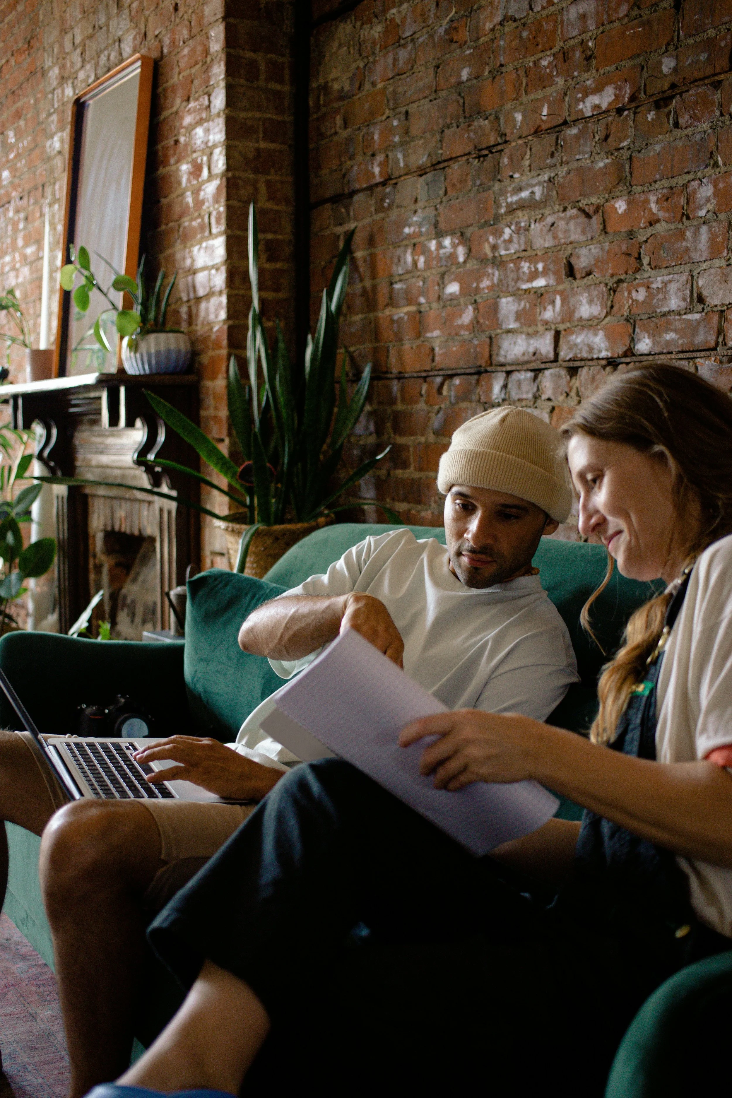 Two young adults are sitting on a green couch in a room with an exposed brick wall. They are looking at papers and a laptop, with plants and a mirror on a shelf in the background.