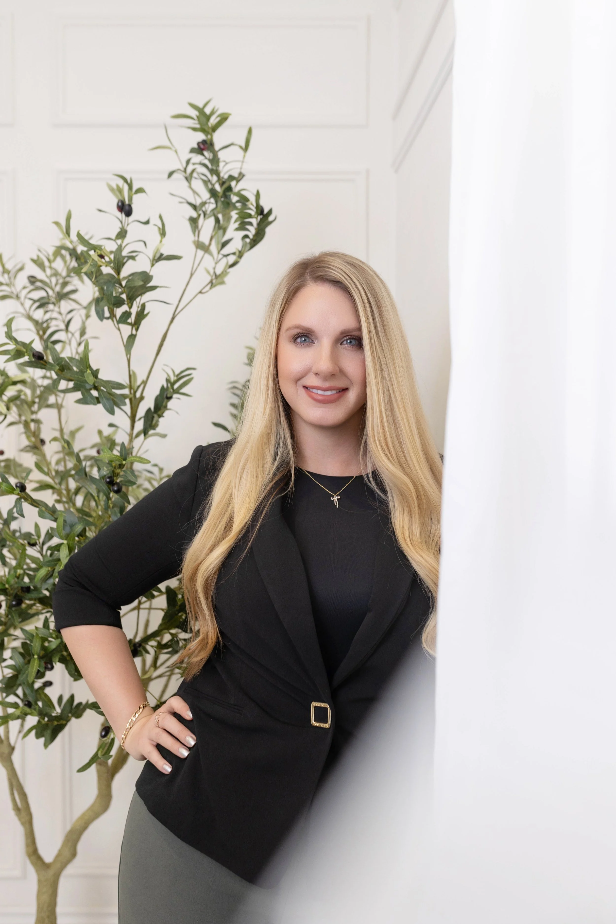 A woman with long blonde hair, wearing a black blazer and a gray skirt, standing indoors next to a green leafy plant with dark berries, with a white paneled wall in the background.