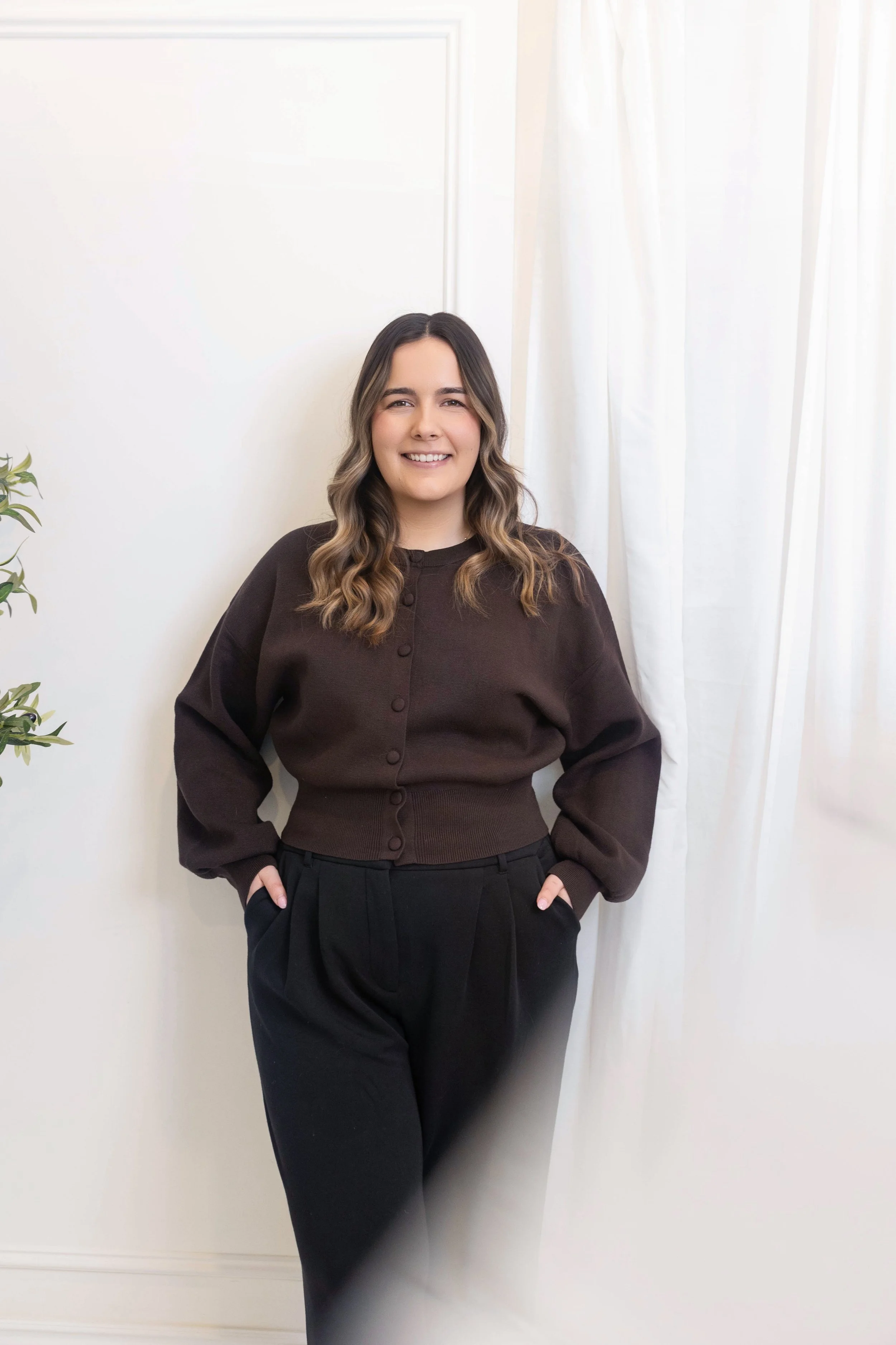 A woman with long wavy hair, wearing a dark brown button-up sweater and black pants, smiling and standing with her hands in her pockets in front of a white wall with a curtain on the right side and a small plant on the left.
