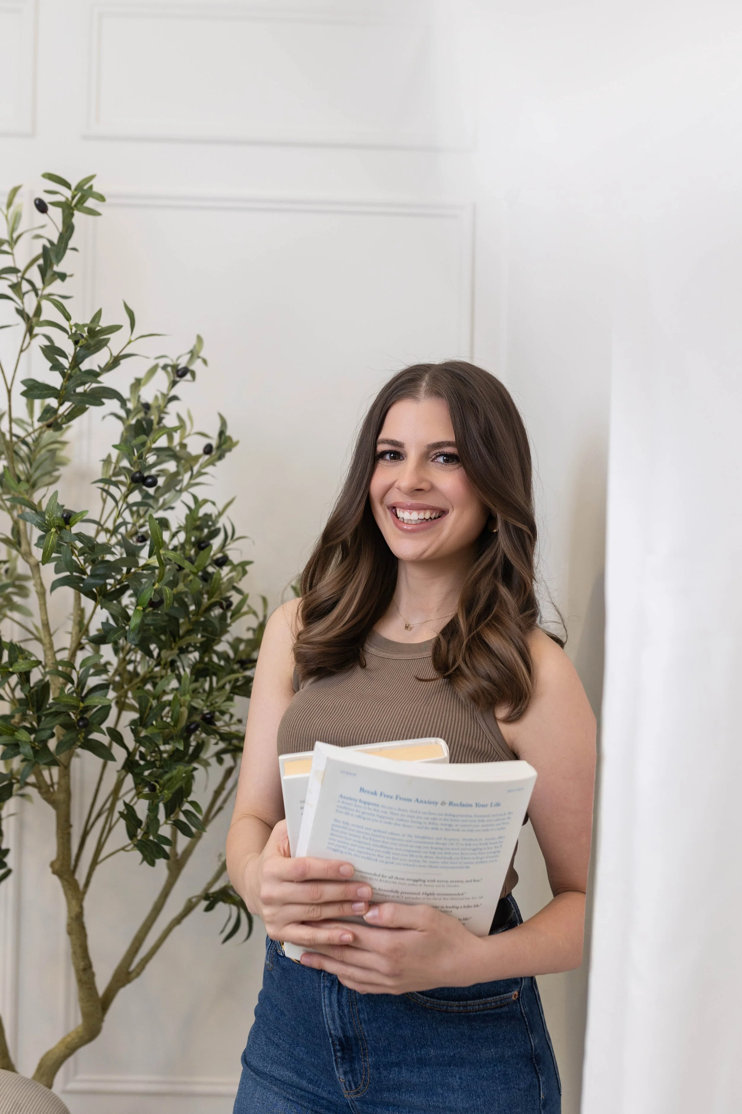 A young woman with long wavy brown hair, smiling and holding a pamphlet, standing next to a potted plant with dark berries, against a white paneled wall.