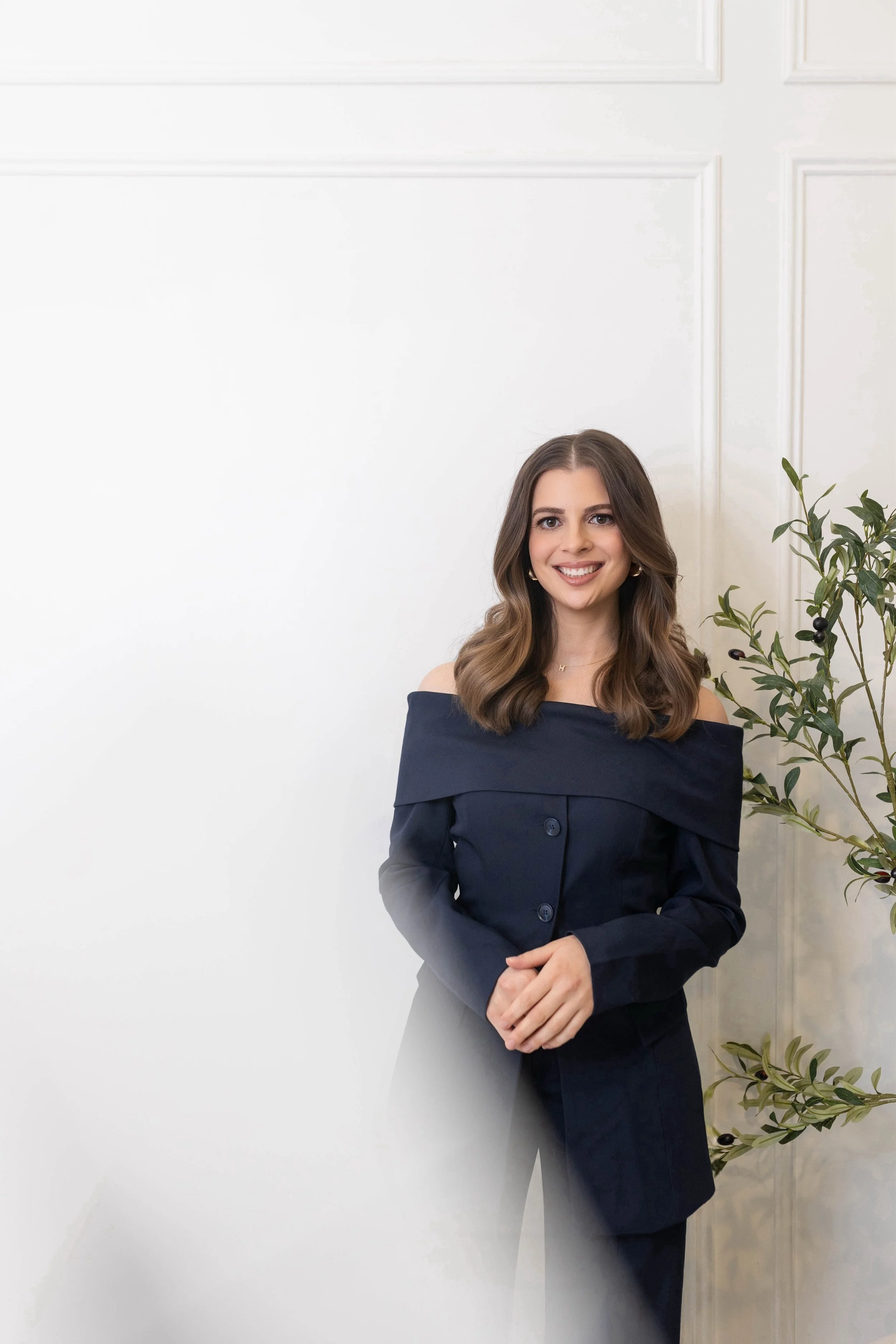A young woman with long wavy brown hair wearing a navy blue off-the-shoulder dress, standing indoors next to a green potted plant with dark purple berries, smiling at the camera.