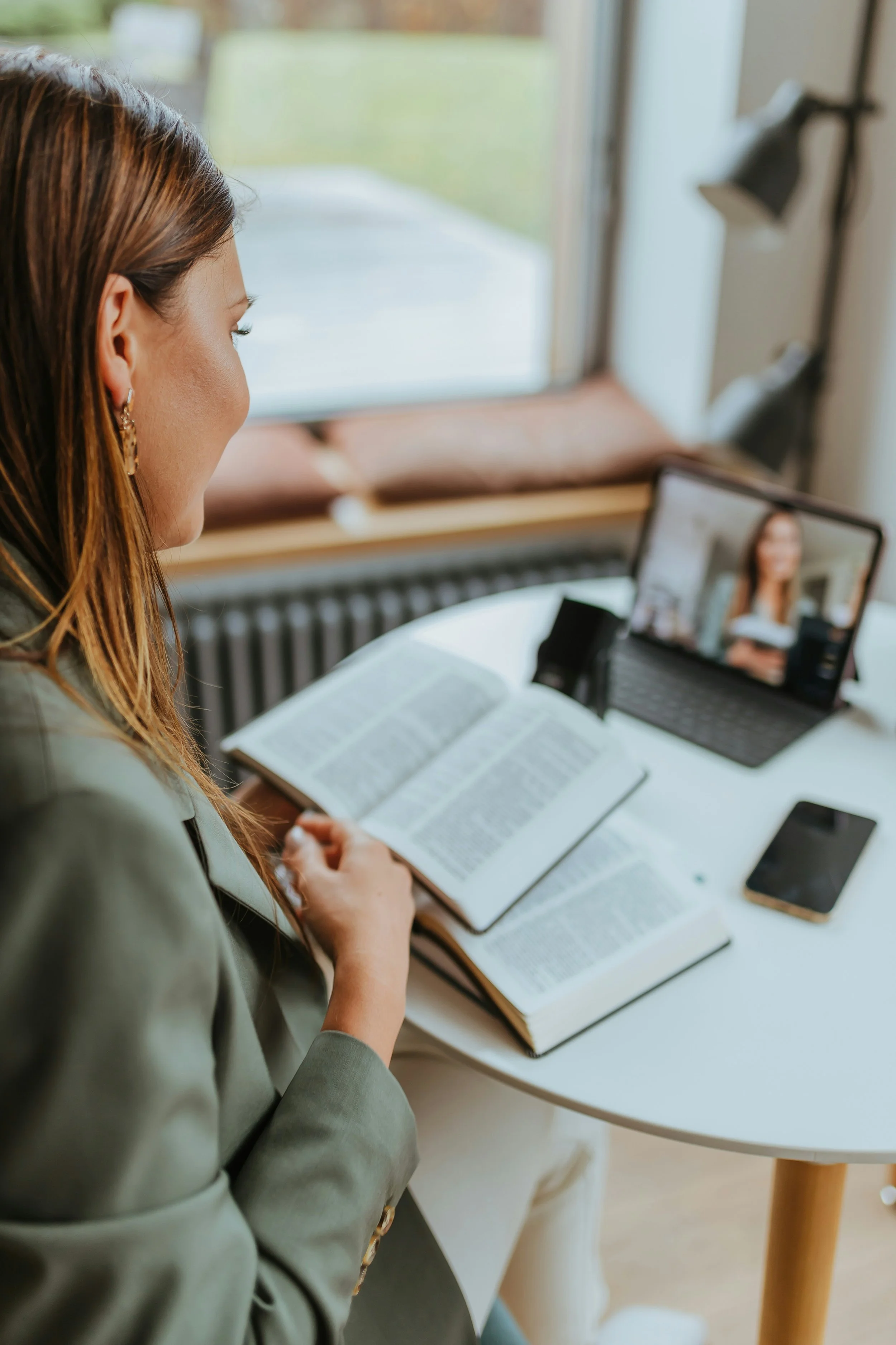 A woman with brown hair wearing earrings, sitting at a table with a book. She is on a video call on a laptop and is holding a cup. There is a smartphone on the table next to an open book. The background shows a window with a view outside and a wall with a lamp.