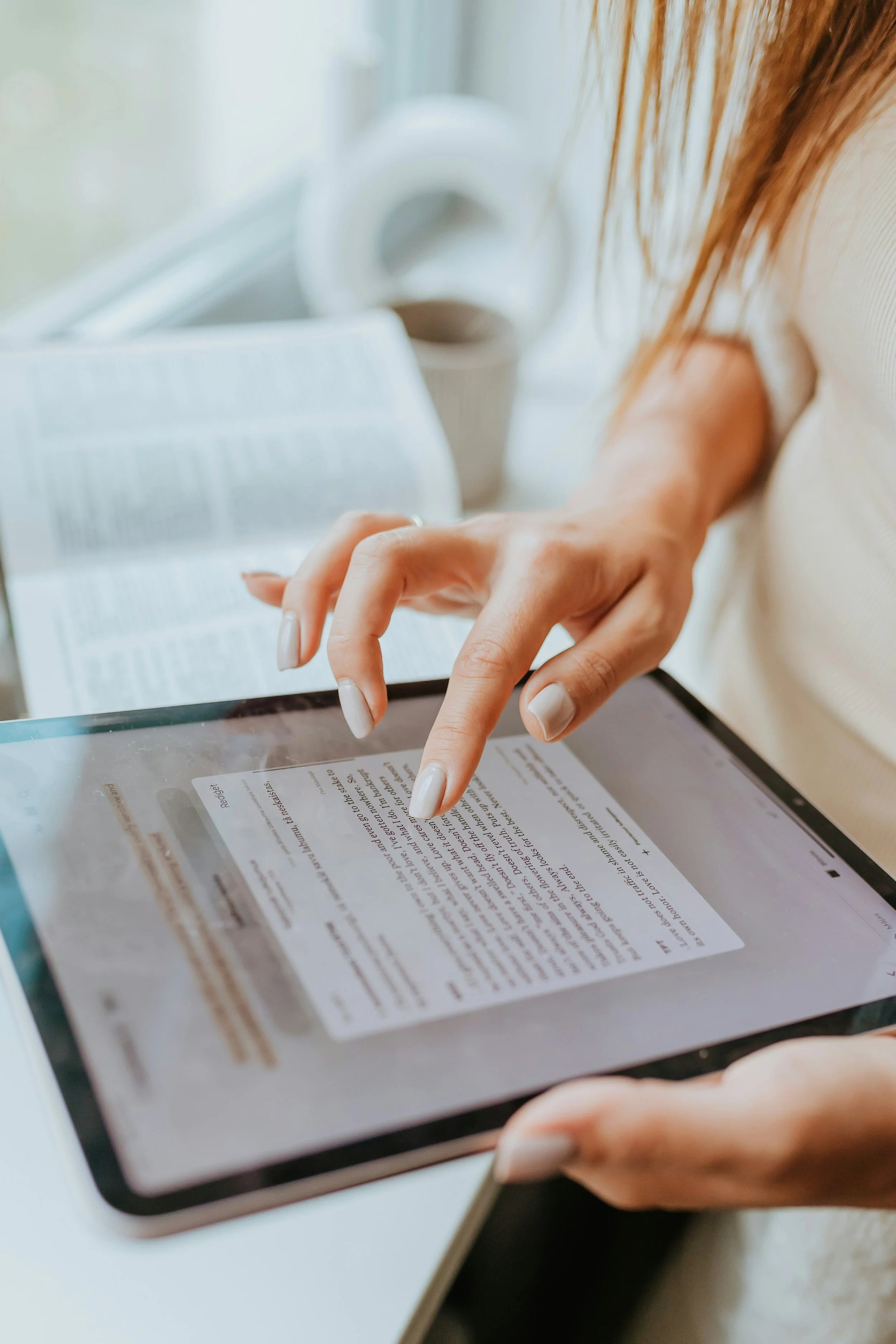 Person using a finger to highlight text on a tablet with an open book in the background.