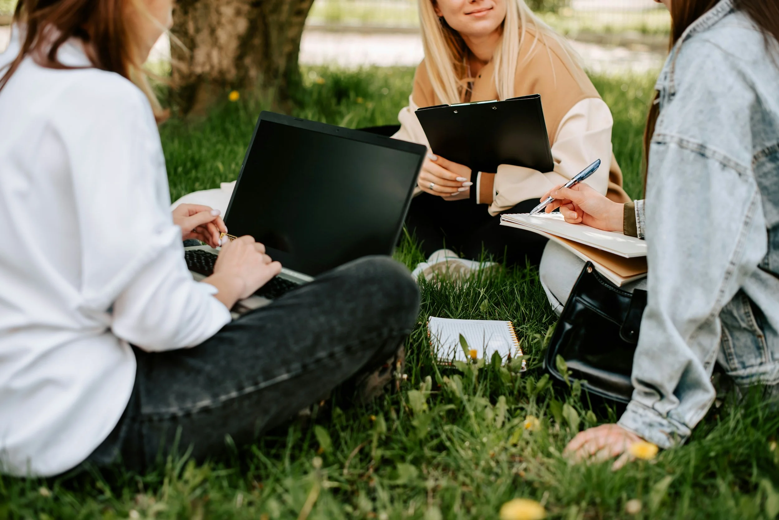 Three women sitting on grass near a tree, holding laptops and notebooks, engaging in outdoor study or work.
