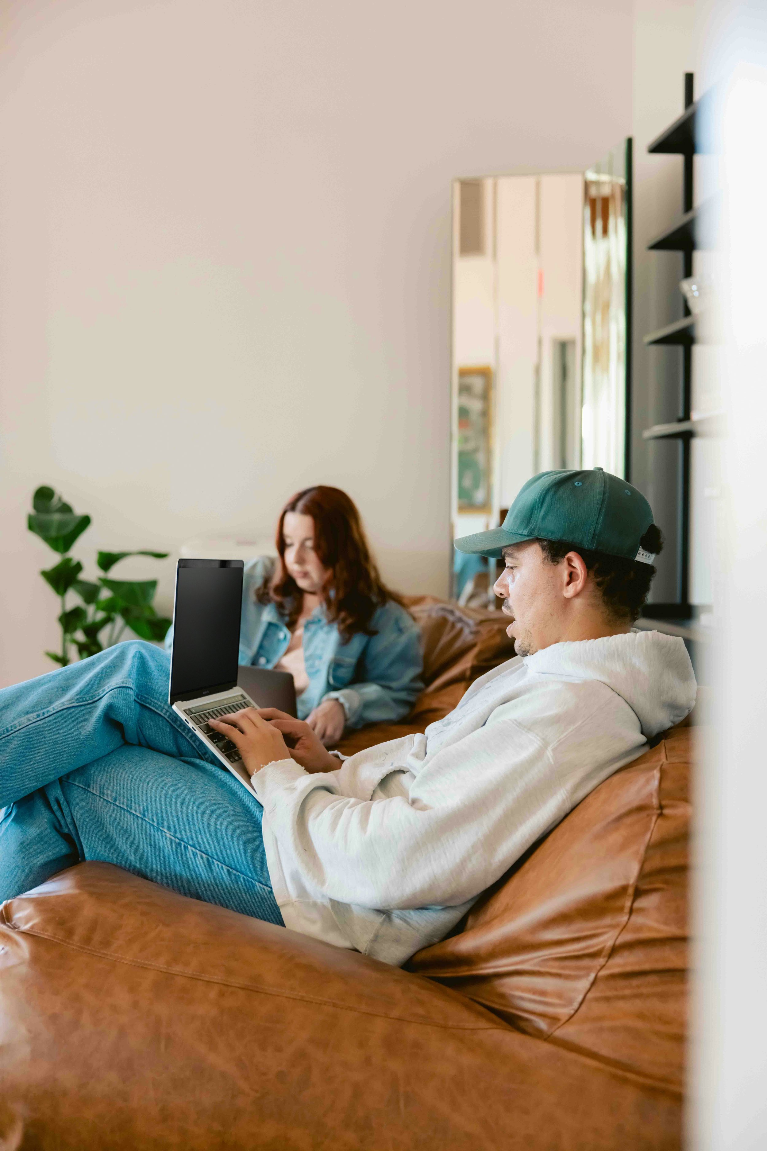 A young man with a baseball cap sitting on a brown leather couch, working on a laptop, with a woman in the background also sitting on the couch and looking at her device. The room is bright with natural light, with plants and shelves on the wall.