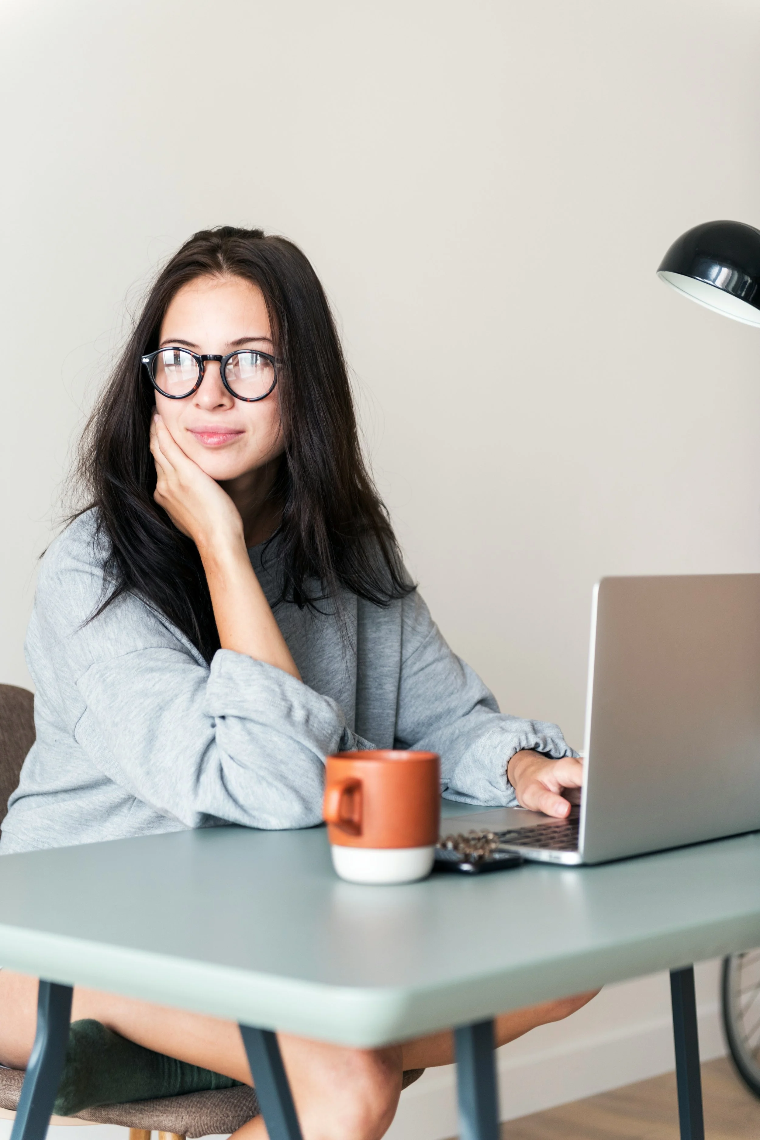 A young woman with long dark hair and glasses sitting at a table with a laptop, a ceramic mug, and some keys, resting her face on her hand and looking thoughtfully at the camera in a minimalist room.