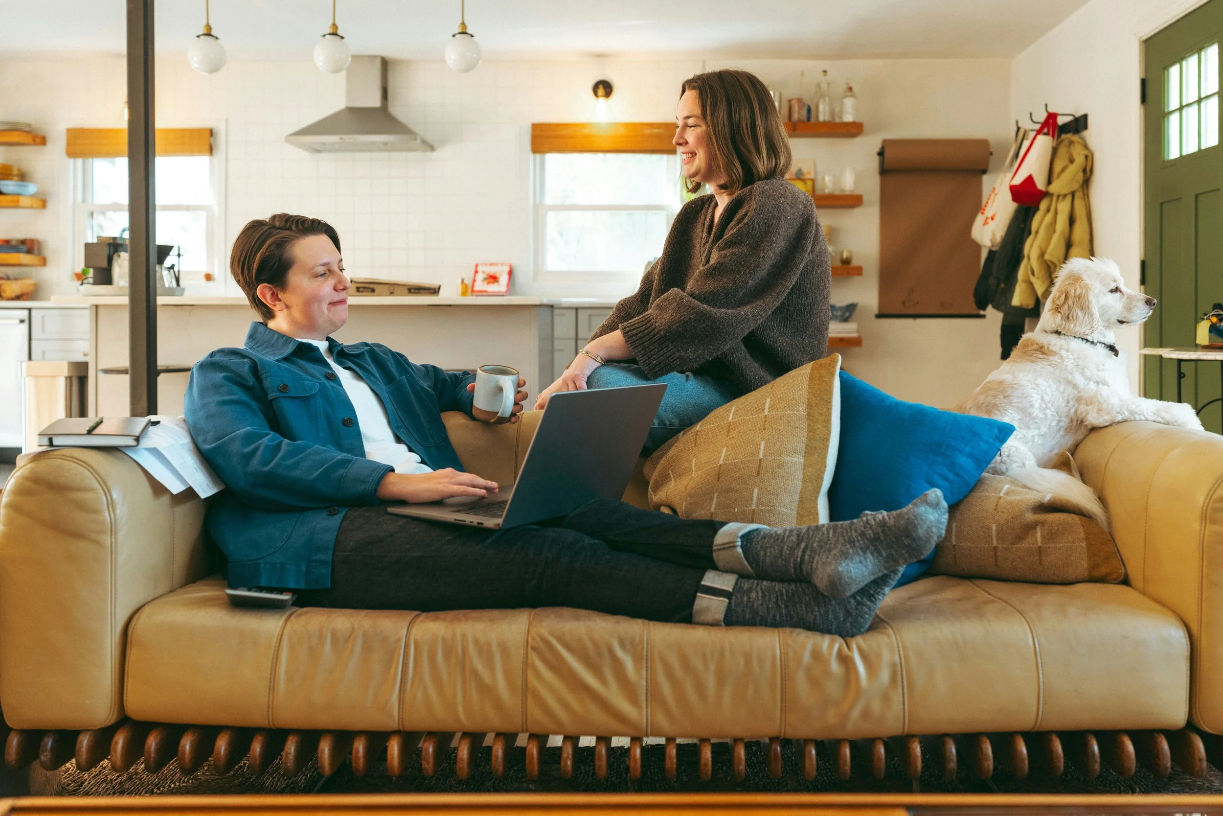 A woman sitting on a yellow leather couch with her feet up, holding a mug, smiling at a woman standing nearby who is leaning on the couch. A golden retriever dog is sitting on the same couch, facing towards the window, with several pillows on the couch. The background shows a kitchen with white cabinets, open shelves, and hanging lights.