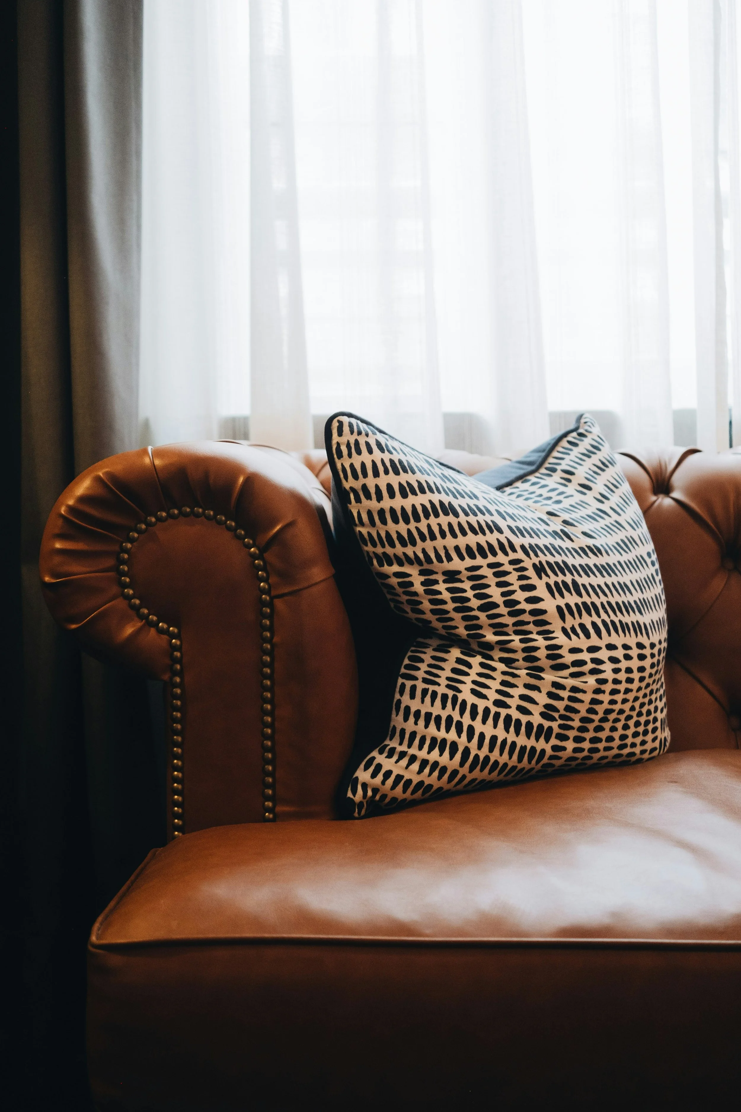 A brown leather sofa with brass nailhead trim and a white and black patterned pillow, positioned in front of a sheer white curtain covering a window.