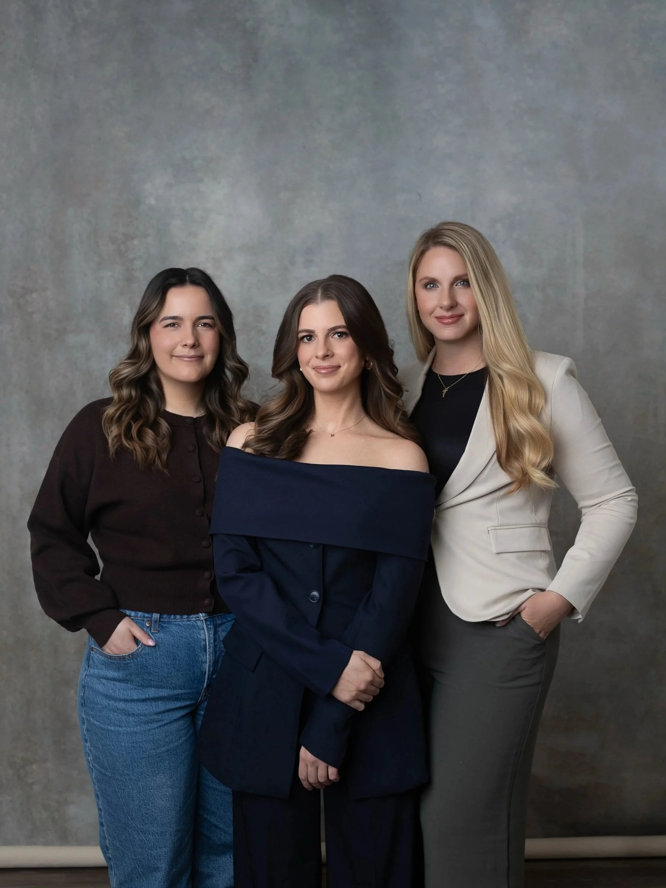 Three women standing close together in front of a textured gray background. The woman on the left has long wavy brown hair, wearing a dark brown button-up shirt and blue jeans. The woman in the center has shoulder-length straight brown hair, wearing a navy blue off-the-shoulder top and matching blazer. The woman on the right has long blonde hair, wearing a black top, a light-colored blazer, and dark gray pants.