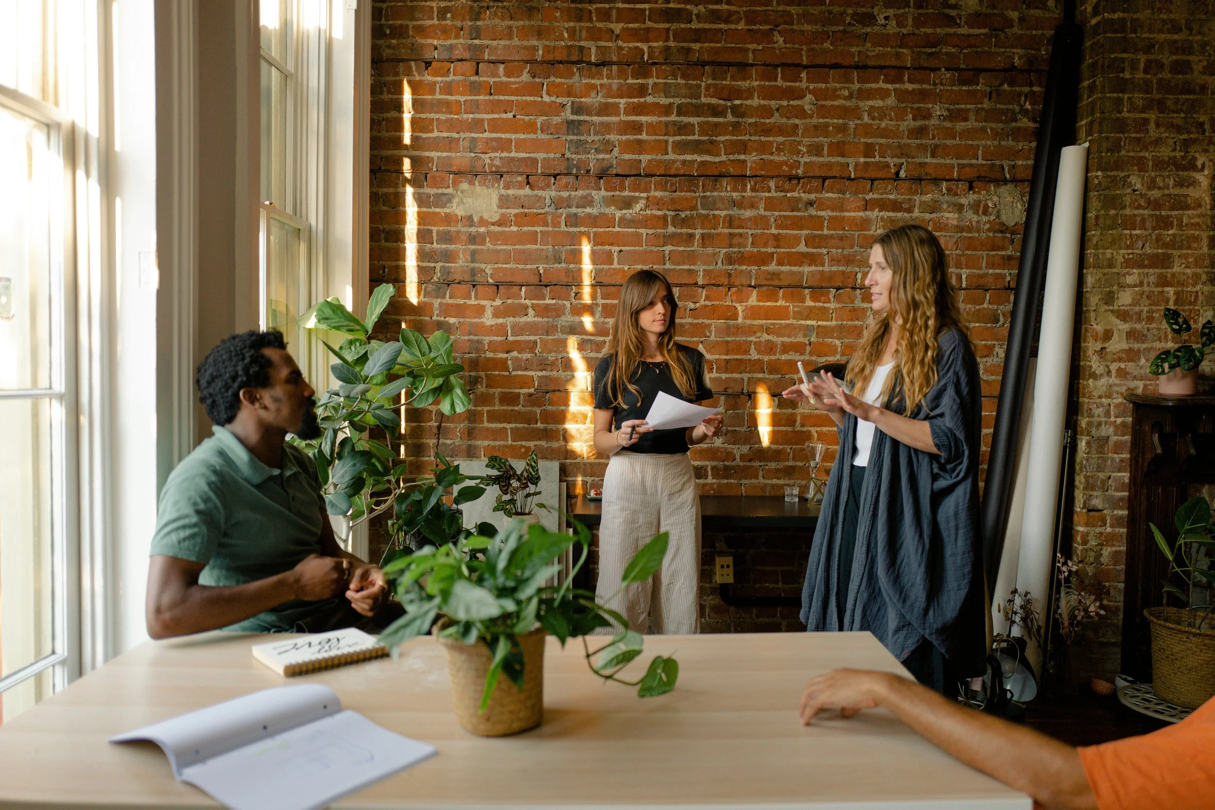 Three women engaged in a discussion in a cozy, well-lit room with an exposed brick wall and large windows, surrounded by plants.