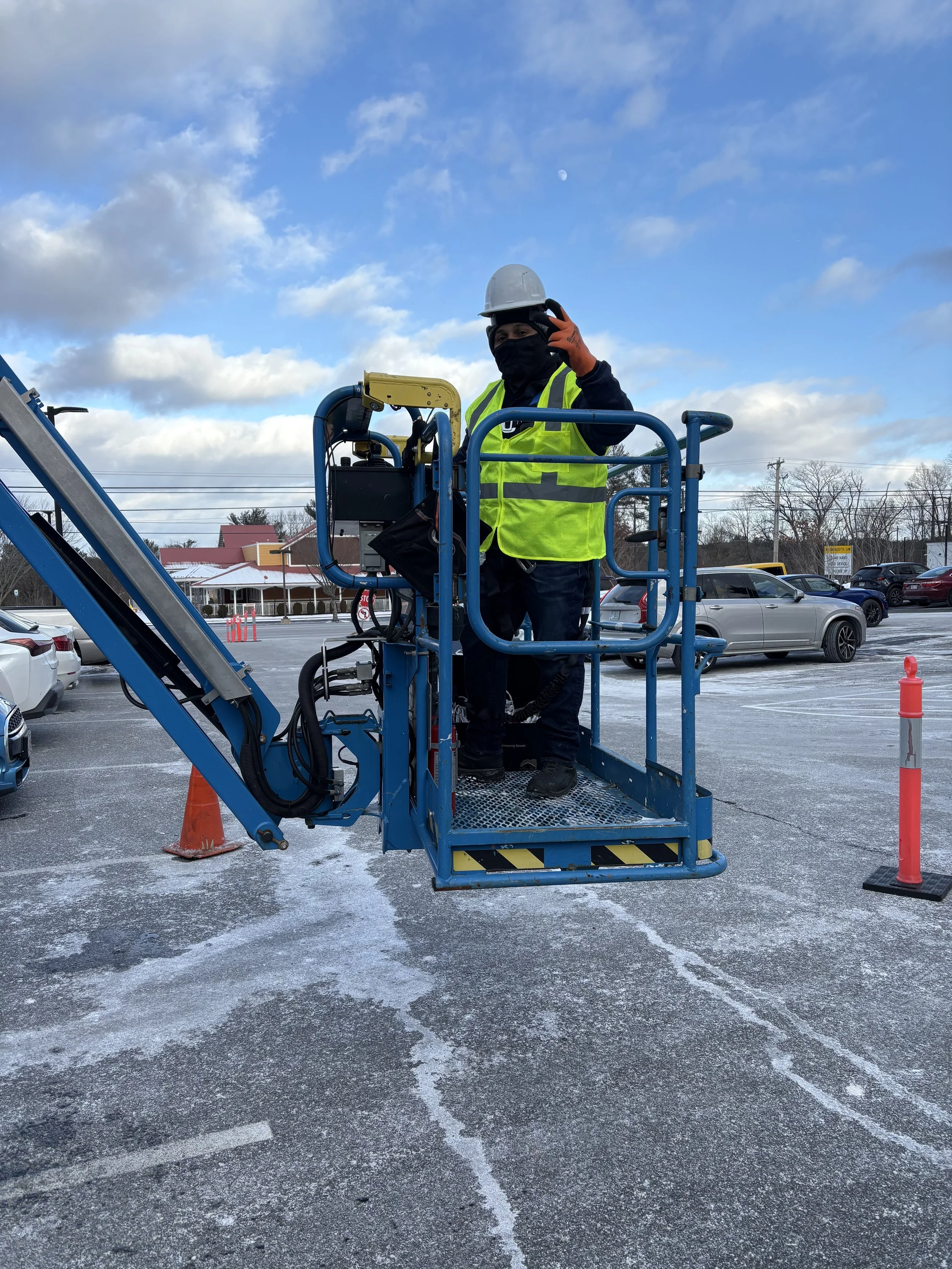 A worker in a safety helmet and a neon yellow reflective vest standing on a blue lift platform in a parking lot, waving at the camera. The parking lot has some parked cars, orange cones, and patches of snow on the ground, with a partly cloudy sky overhead.