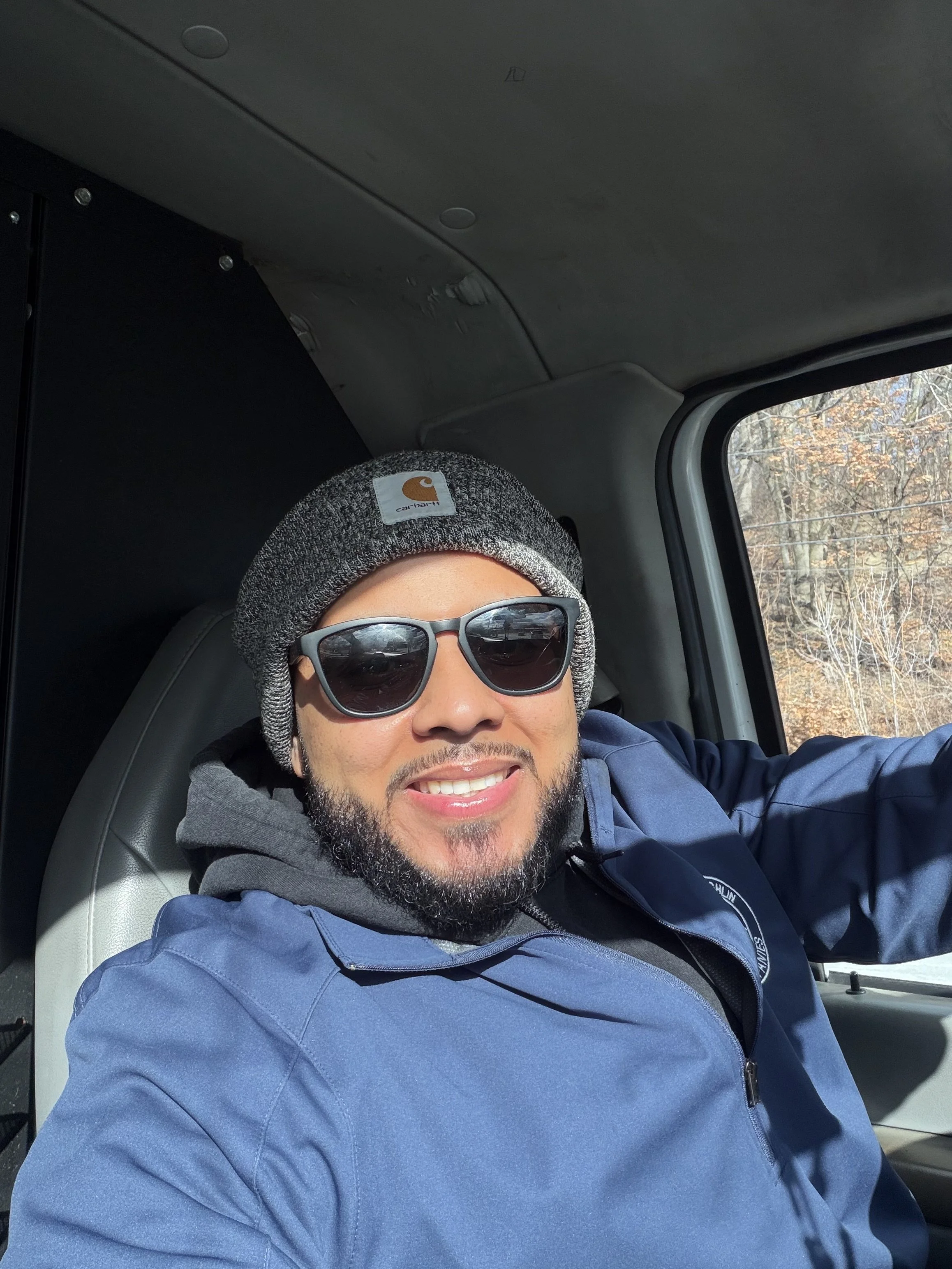 A man with a beard and sunglasses wearing a gray beanie and blue jacket, smiling while sitting in a vehicle with trees visible outside the window.