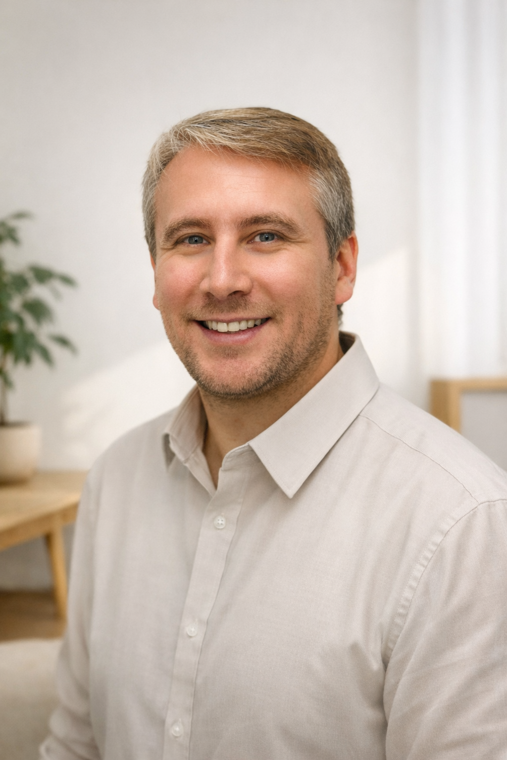 Portrait of a smiling man with short light brown hair, wearing a light beige button-up shirt, in a bright indoor setting with a blurred background including a potted plant and furniture.