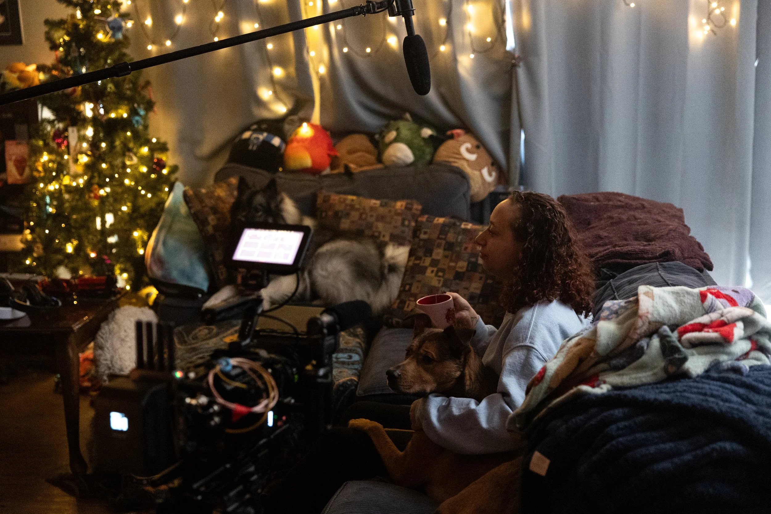 A woman with curly hair holds a mug while sitting on a couch, with a dog nearby, in a cozy living room decorated for Christmas with a lit Christmas tree, plush pillows, stuffed animals, and string lights.