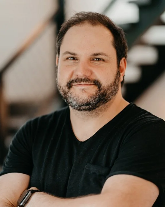 A man with dark hair, beard, and mustache, wearing a black t-shirt, smiling and crossing his arms in front of a staircase in an indoor setting.