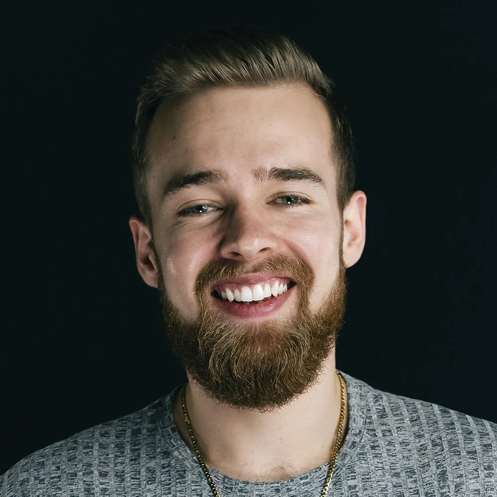 A smiling man with a beard and mustache, wearing a gray textured shirt and gold chain necklace, against a black background.