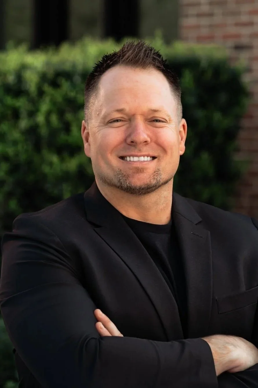 A man with short dark hair, a goatee, wearing a black blazer over a black shirt, smiling with arms crossed, outdoors with green bushes and a brick wall in the background.