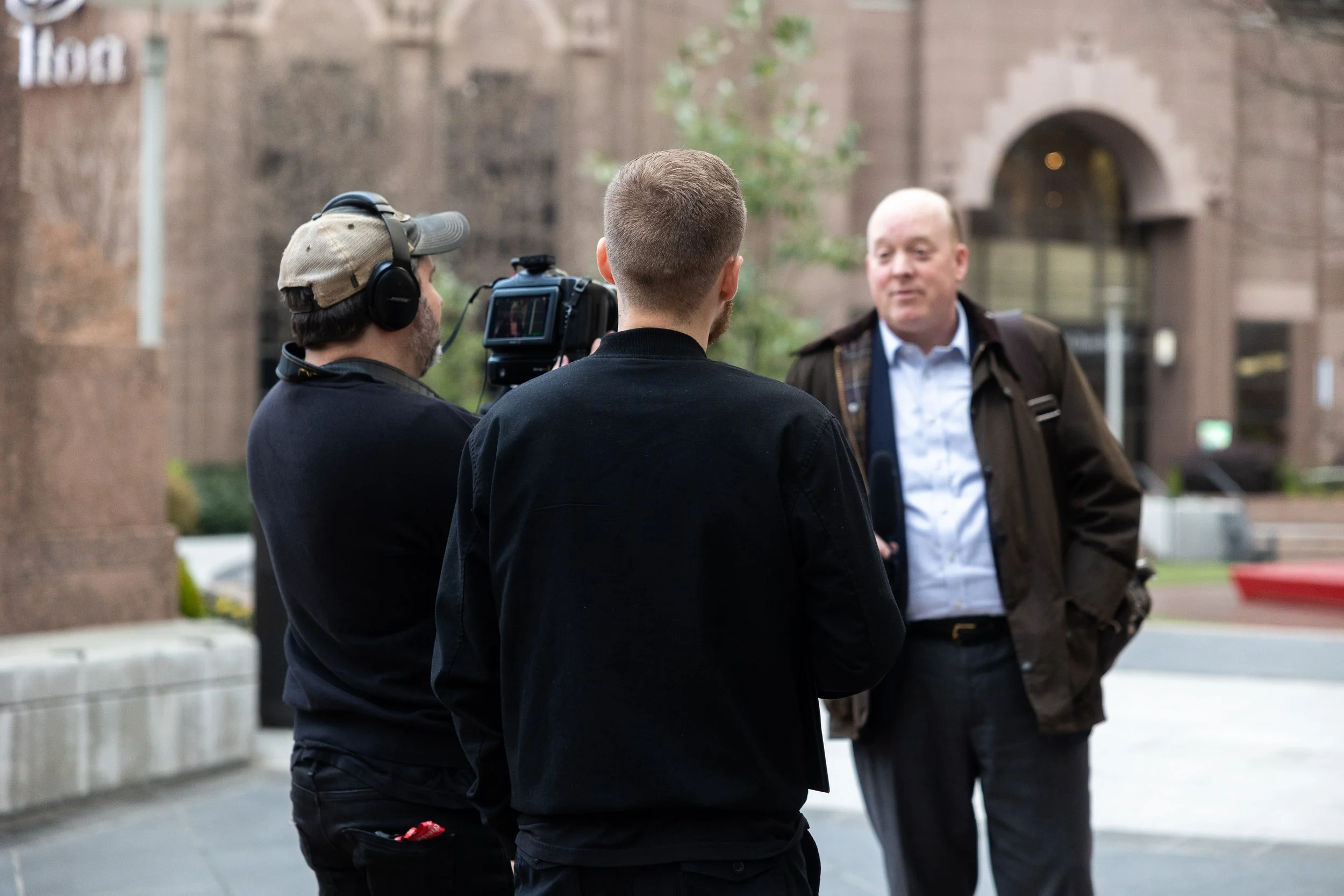 Two men filming a conversation with a third man on a city street, with brick buildings in the background.