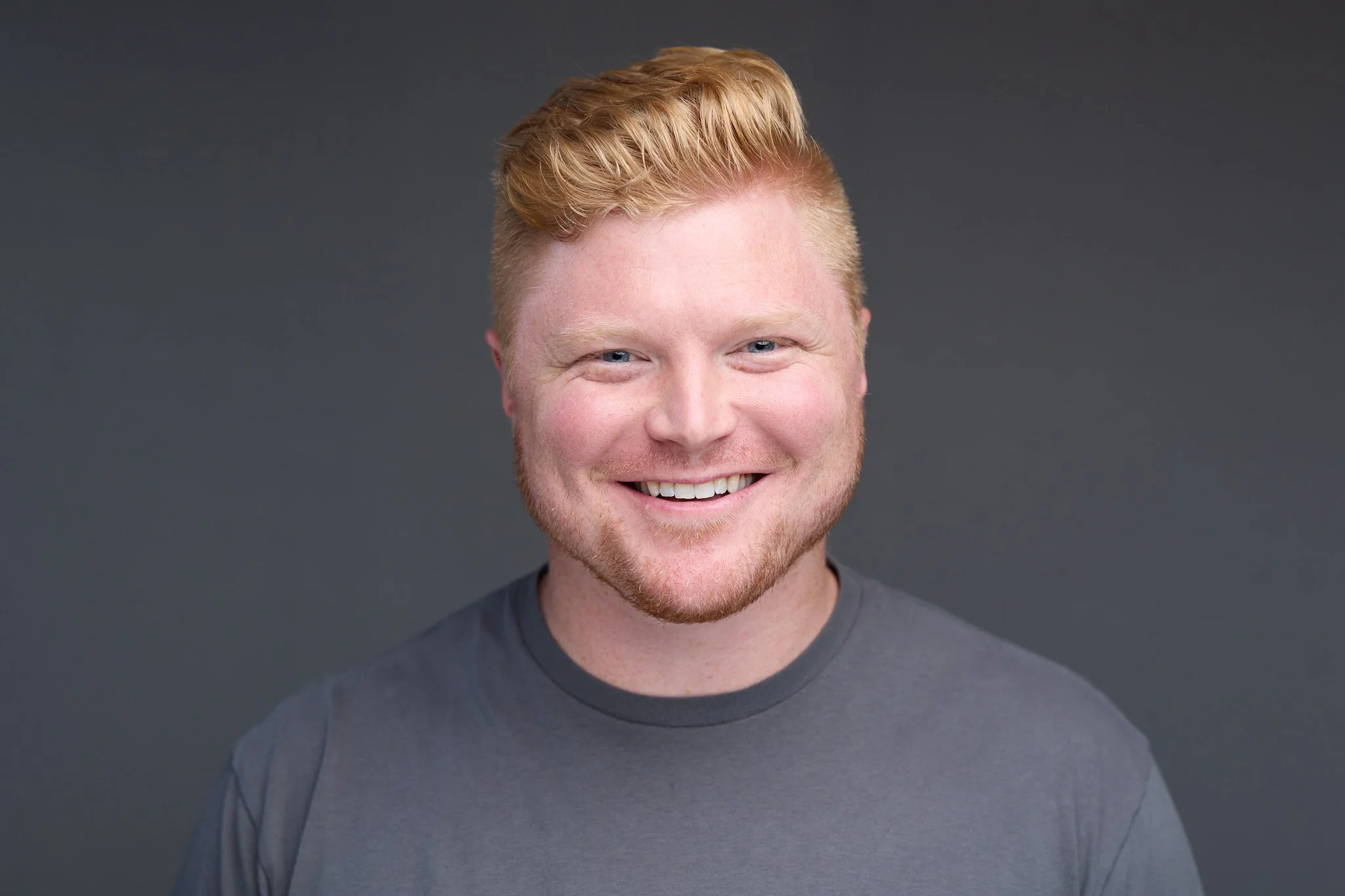 A smiling man with ginger hair and a beard, wearing a gray t-shirt, against a dark gray background.