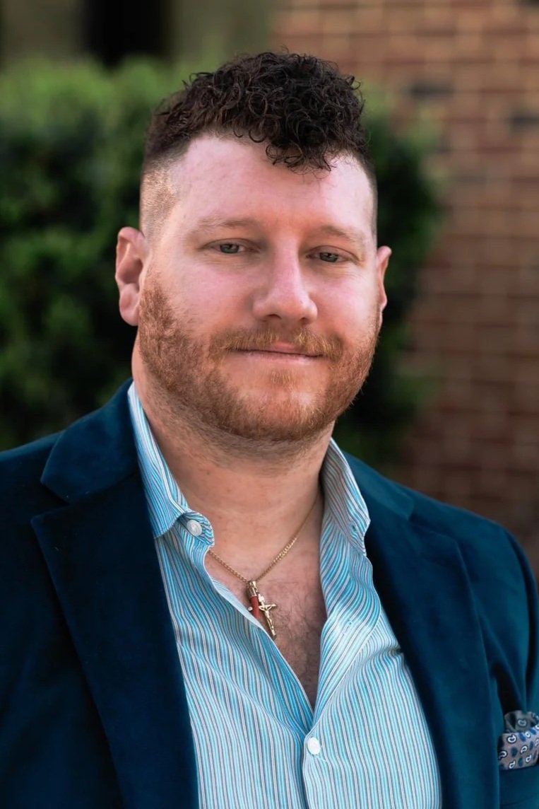 A man with curly dark hair, a beard, and light skin, wearing a blue suit jacket over a blue and white striped shirt, with a gold chain and cross necklace, standing outdoors with greenery and a brick wall in the background.