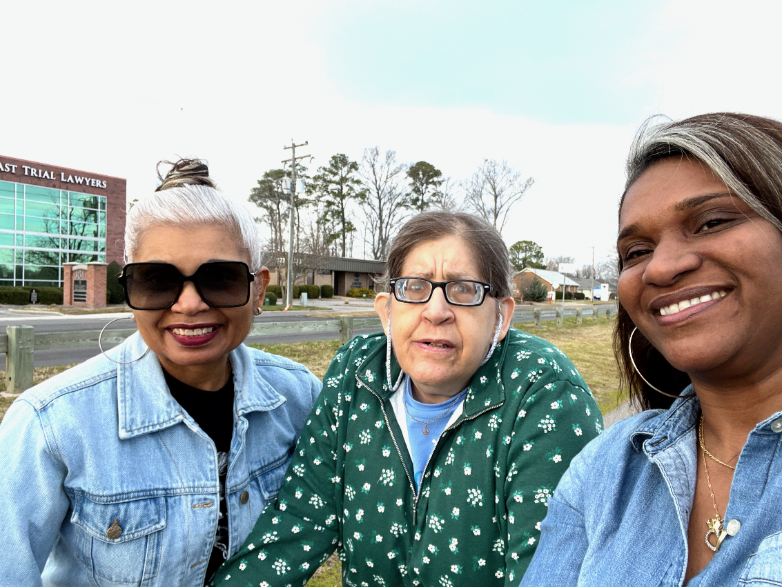 Three women taking a selfie outdoors with a building and trees in the background, one woman wearing sunglasses, another with glasses and a green jacket, and the third smiling with earrings and a necklace.