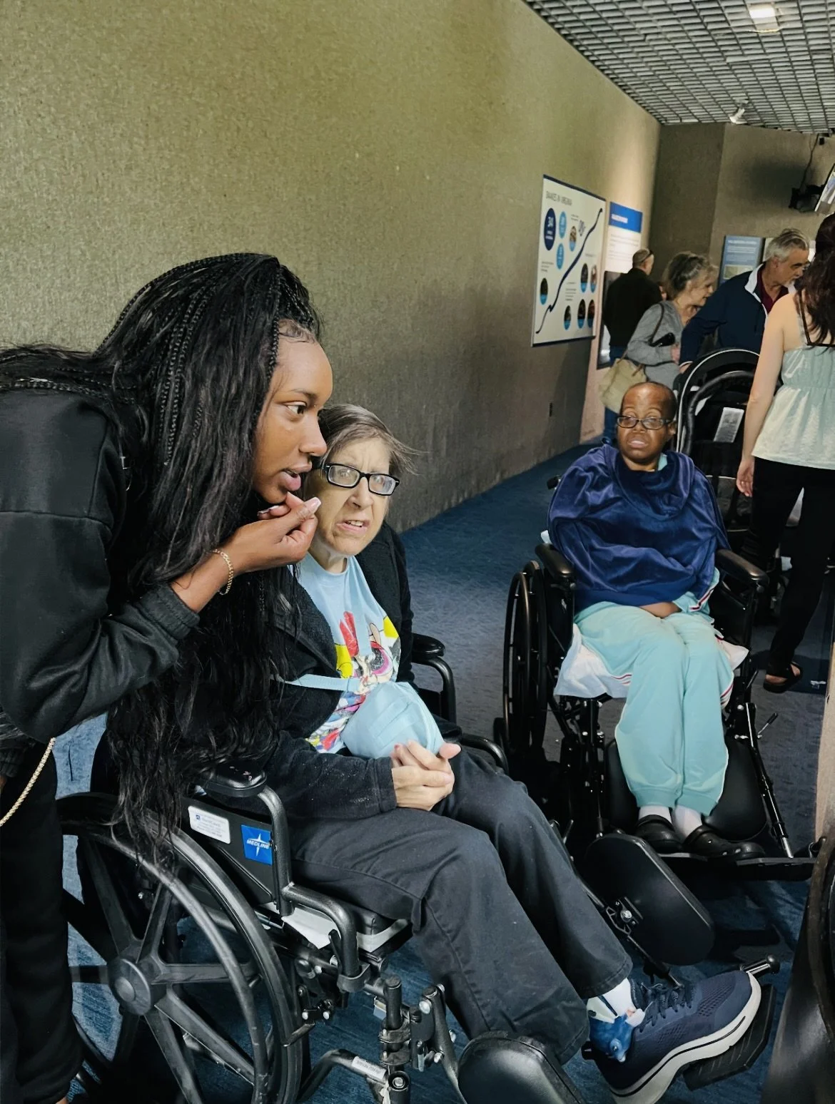 People in wheelchairs at an indoor public space, two women having a conversation, background with informational posters and other visitors.
