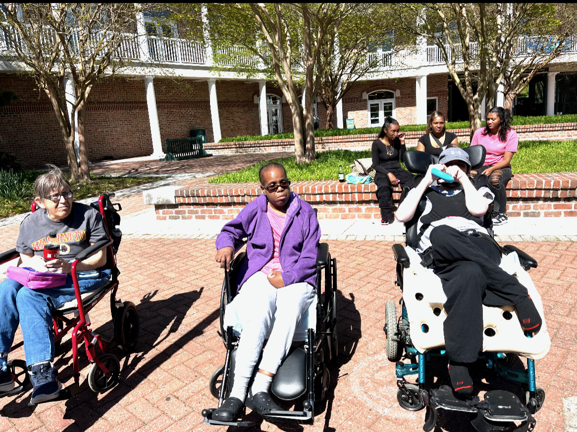 Group of people outdoors sitting on brick ledges and in wheelchairs, with trees and a brick building in the background.