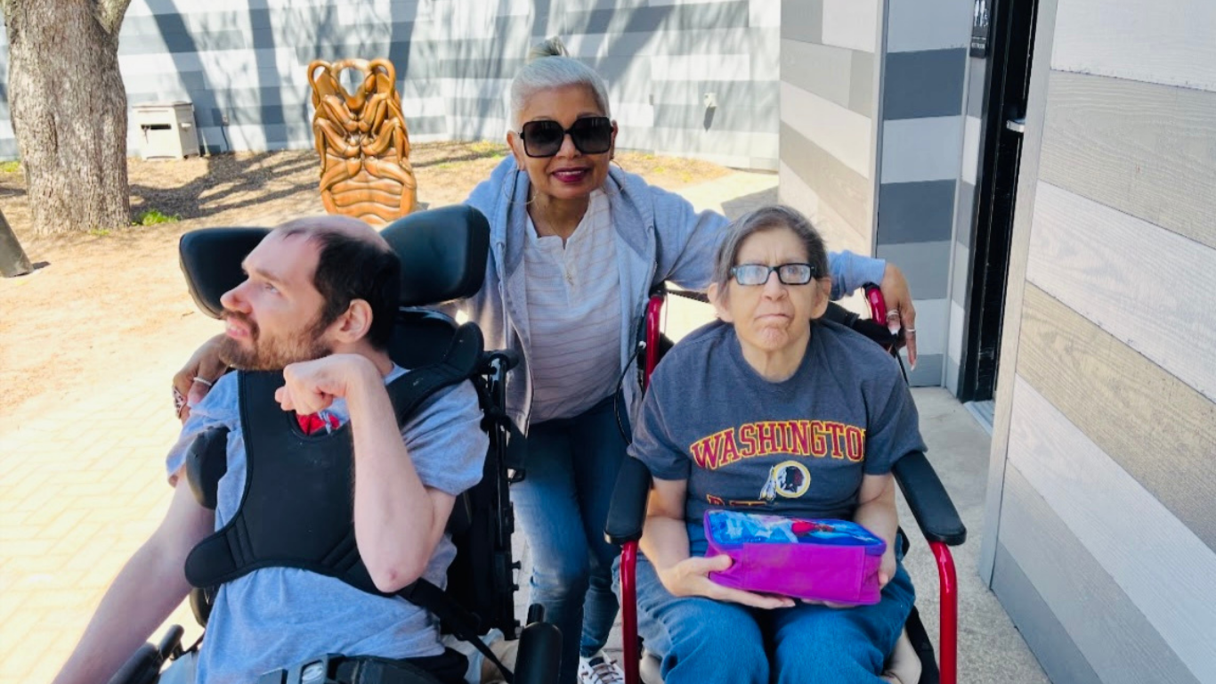 Two elderly women in wheelchairs outdoor, one smiling at the camera and the other holding a wrapped gift, with a caregiver standing behind them.