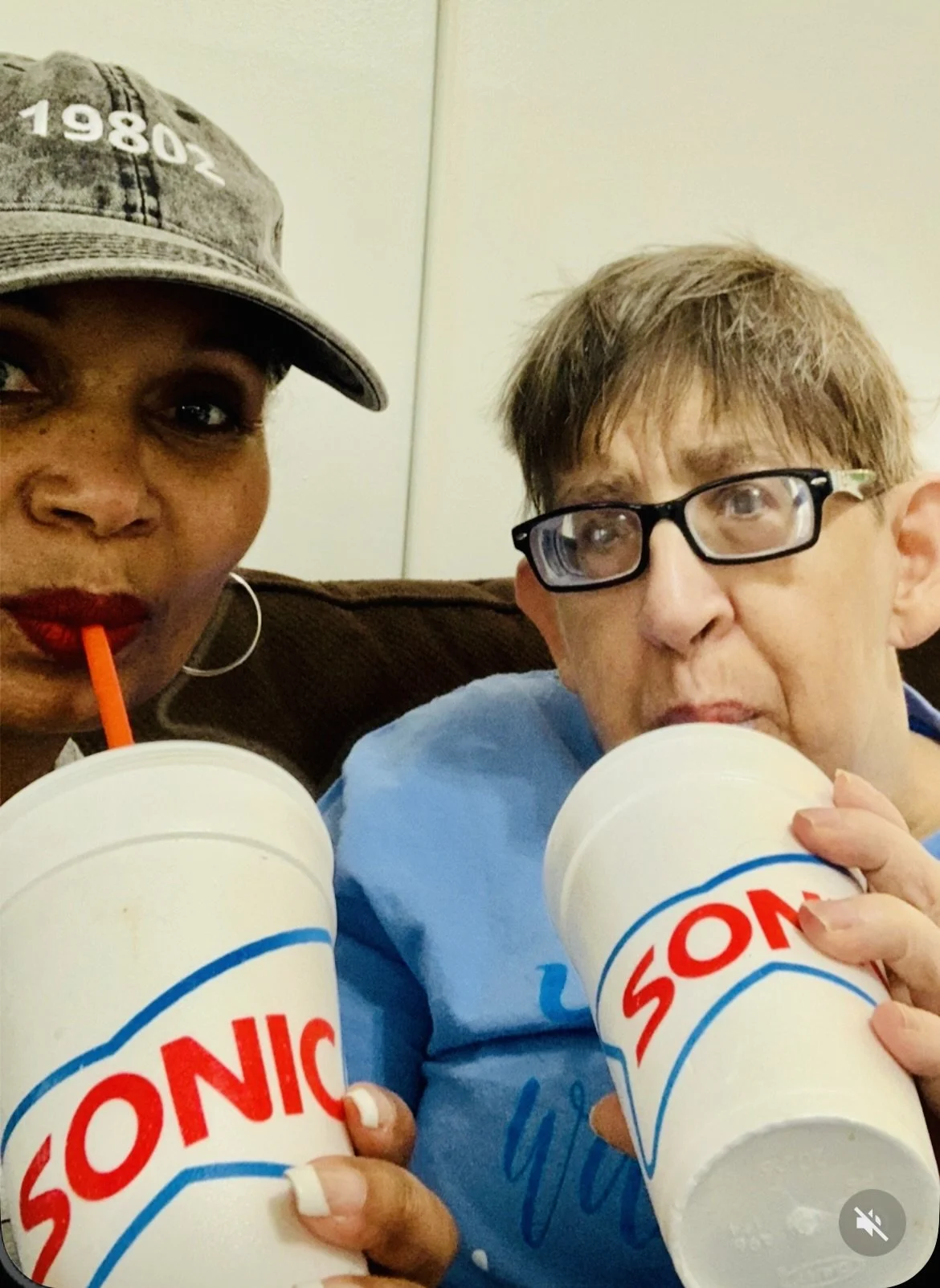 Two people sitting on a couch, each holding a Sonic Drive-In cup with straws, enjoying a drink. The woman on the left is wearing a gray cap and red lipstick. The person on the right has short brown hair and glasses.