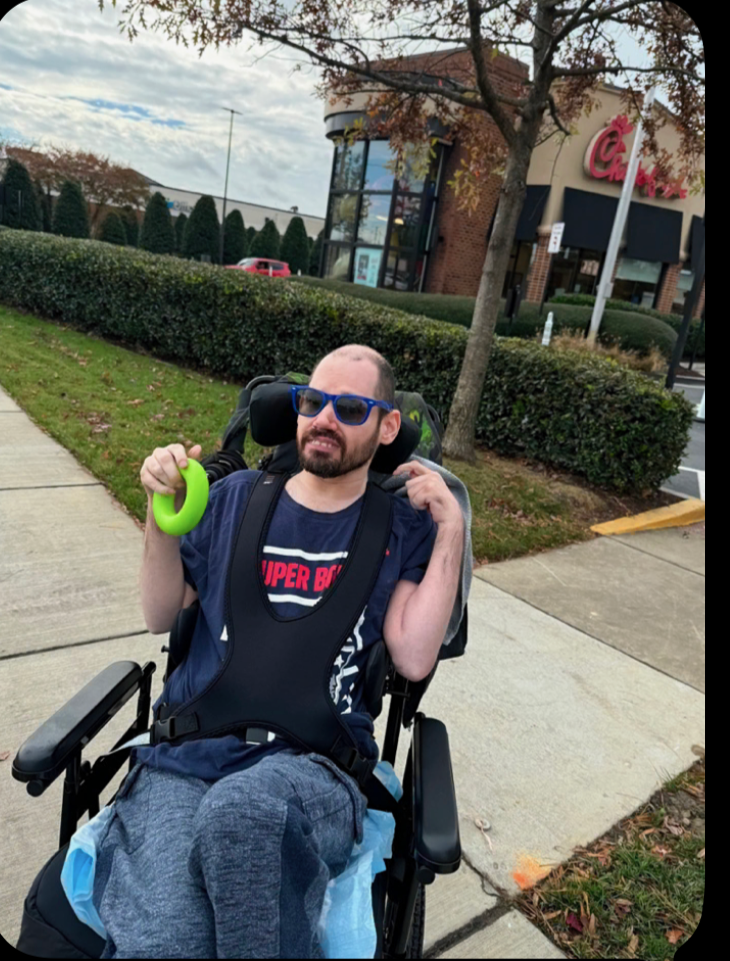 A man with a beard wearing sunglasses, a dark blue shirt, and gray sweatpants sitting in a motorized wheelchair outside a shopping plaza. He is holding a green ring in his right hand and resting his left hand on his shoulder strap, with a Chick-fil-A