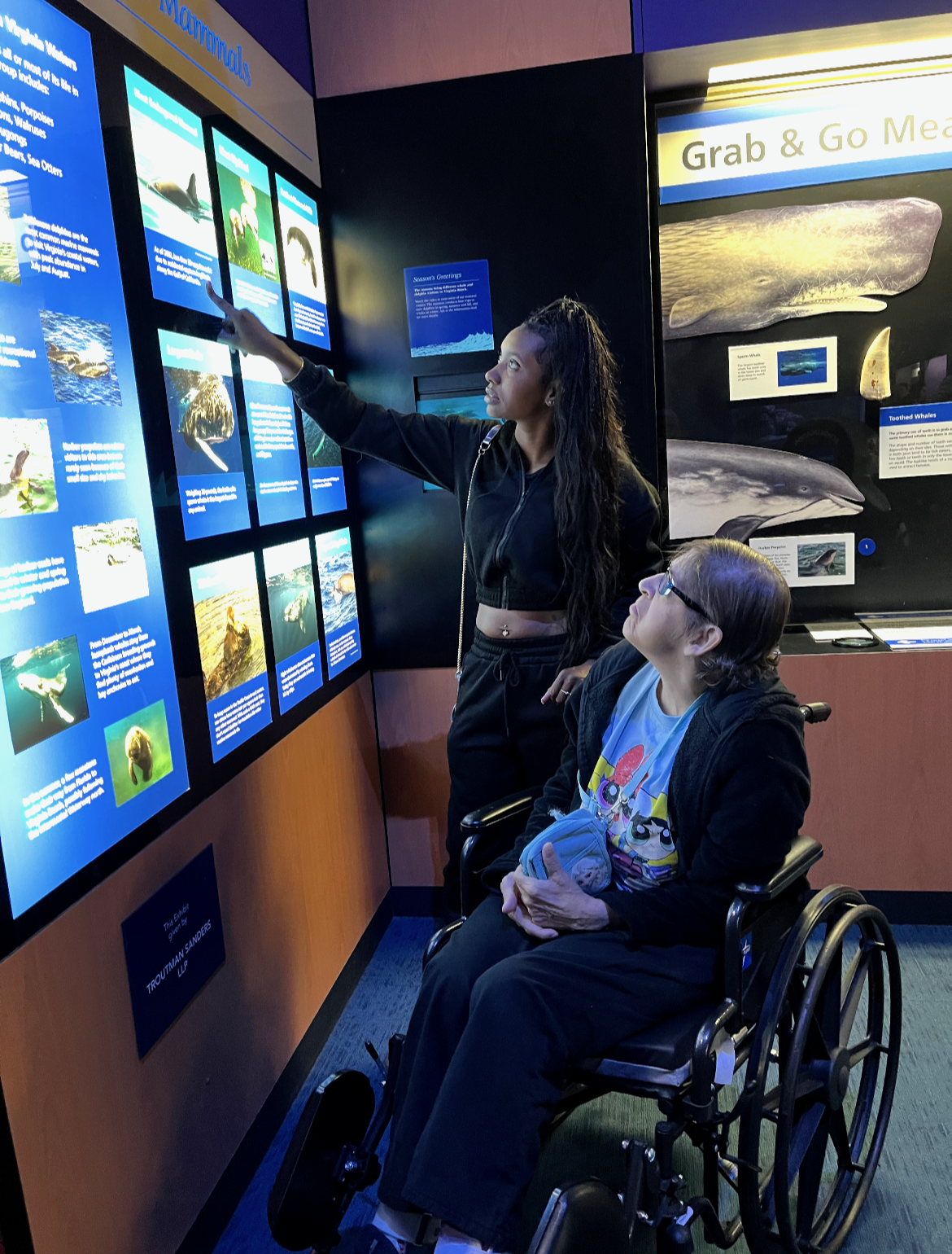 Two women, one in a wheelchair, looking at an educational display about marine life at a museum. The woman in the black hoodie is pointing at information on the display, while the other woman looks on.