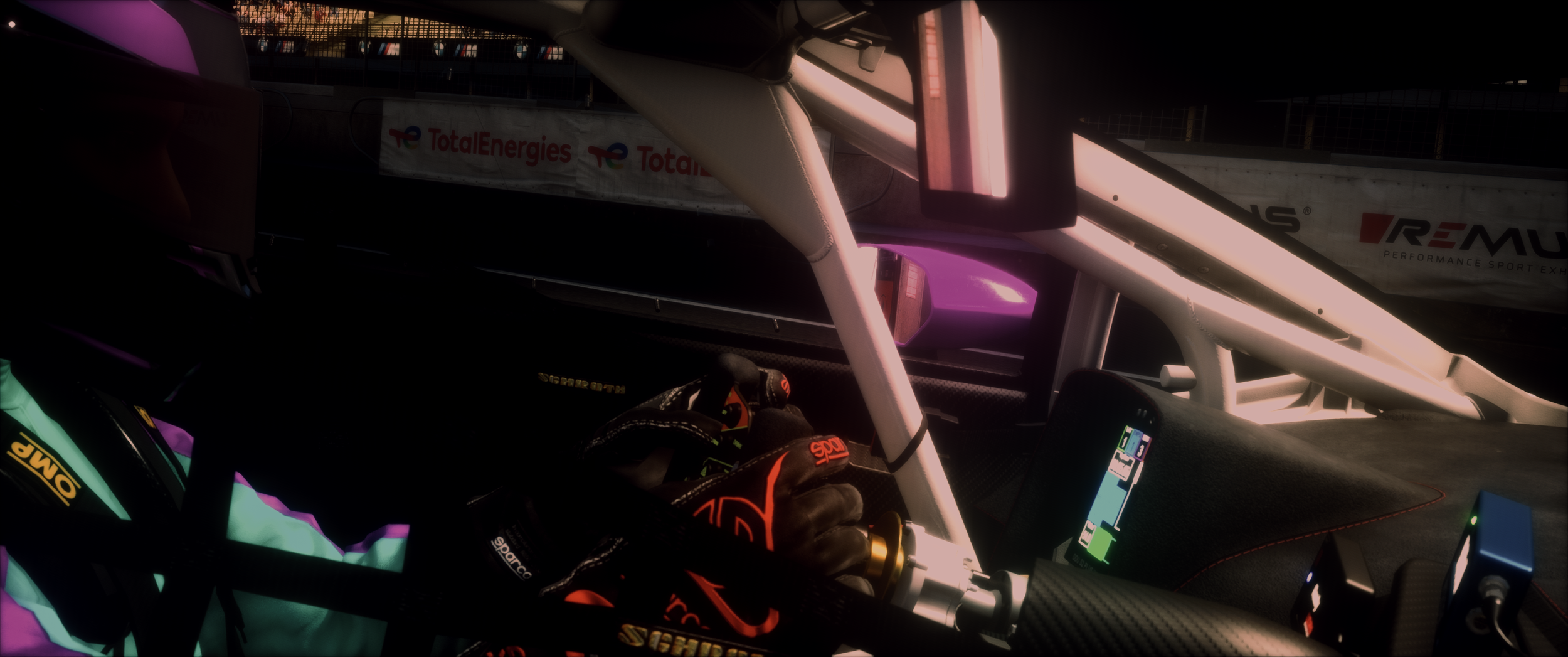 Inside view of a race car cockpit showing a driver wearing racing gloves, steering wheel, digital display, and part of the roll cage. The background features advertising banners.