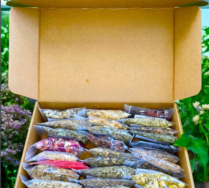 Open cardboard box filled with sealed plastic bags of various dried herbs and plant materials, with a green garden background.