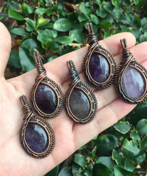 A hand holding five wire-wrapped pendants with oval dark purple stones against a green leafy background.