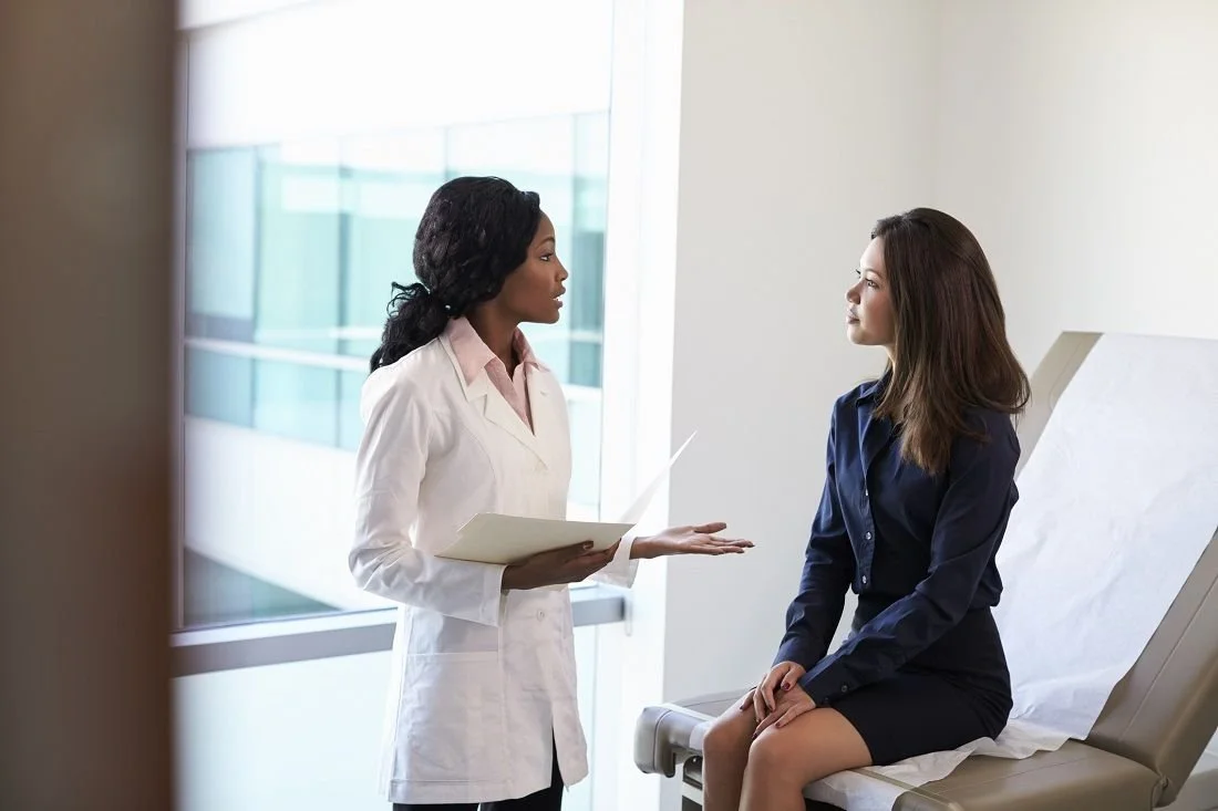 A doctor and a woman having a discussion in a medical office, with the woman sitting on an examination table.