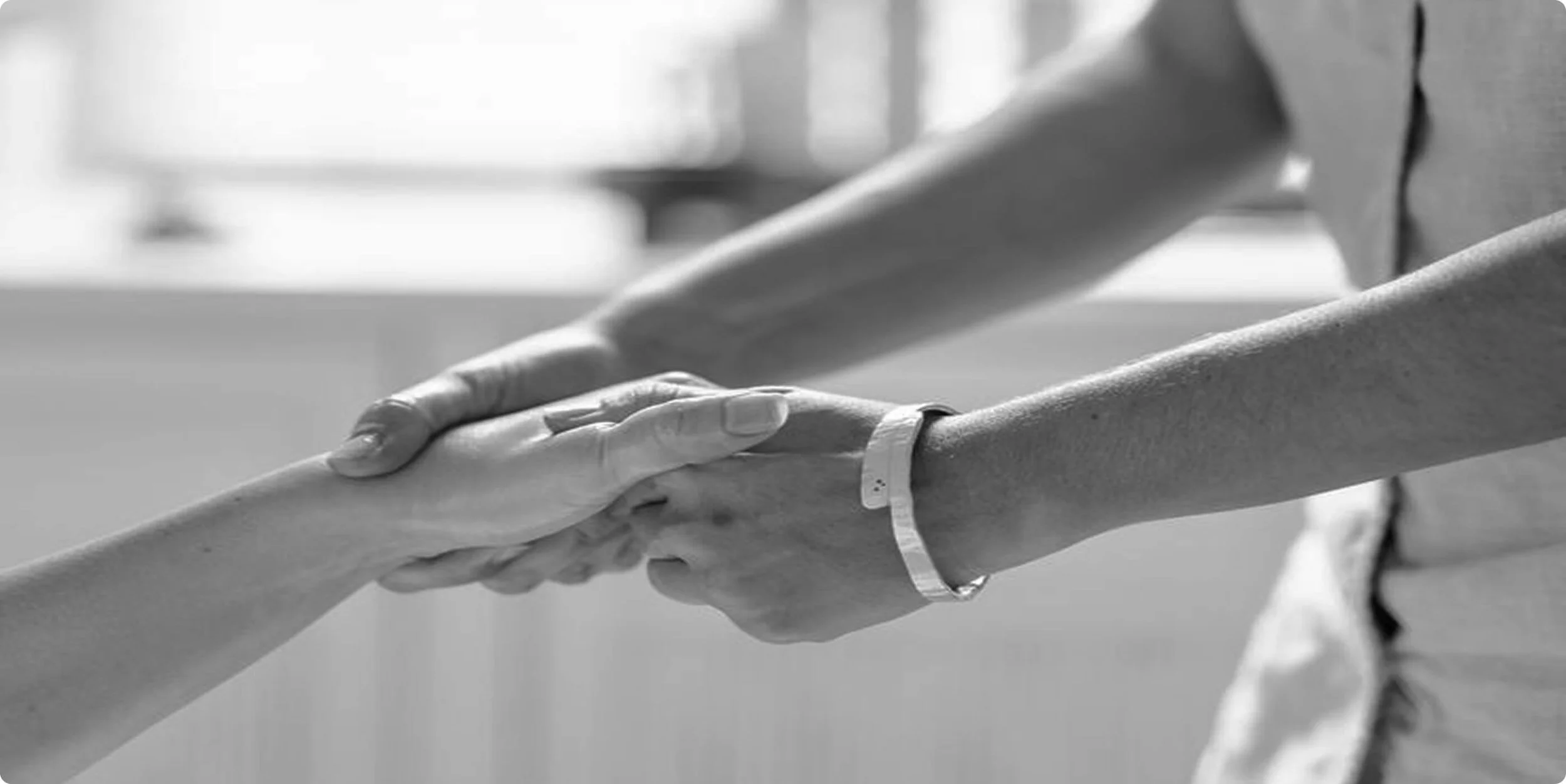 A close-up black and white photo of two hands holding each other, one wearing a hospital bracelet.
