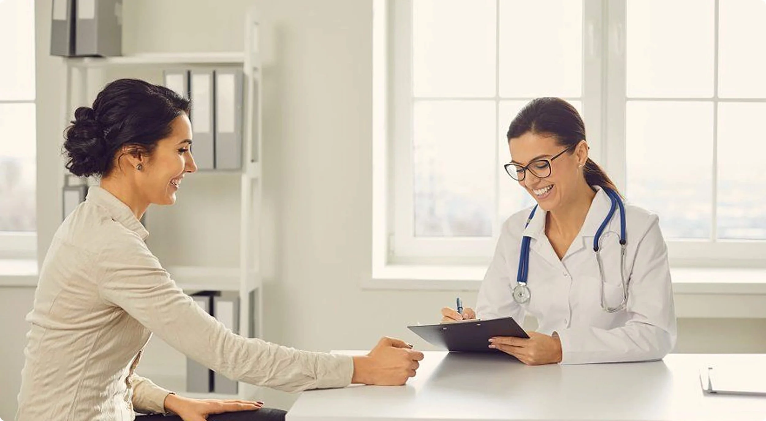 A woman visiting a doctor in a medical office, talking and smiling with the female doctor who is wearing a white coat and stethoscope and holding a tablet.
