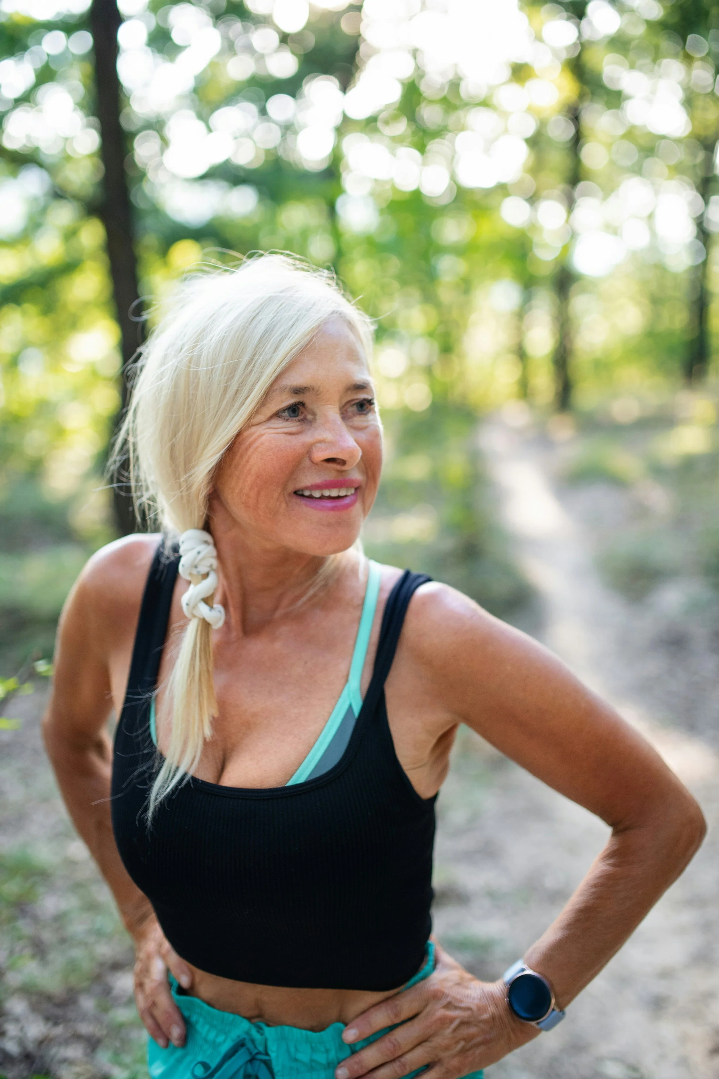 A smiling senior woman with long blonde hair in a braid, wearing a black tank top, turquoise sports bra, turquoise shorts, and a smartwatch, standing outdoors on a forest trail with lush greenery and sunlight filtering through the trees.