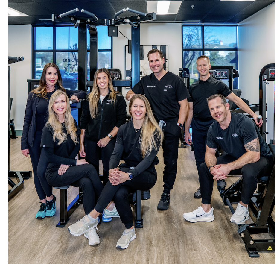 Group of six people, four women and two men, smiling inside a gym or fitness center, with workout equipment and large windows in the background.