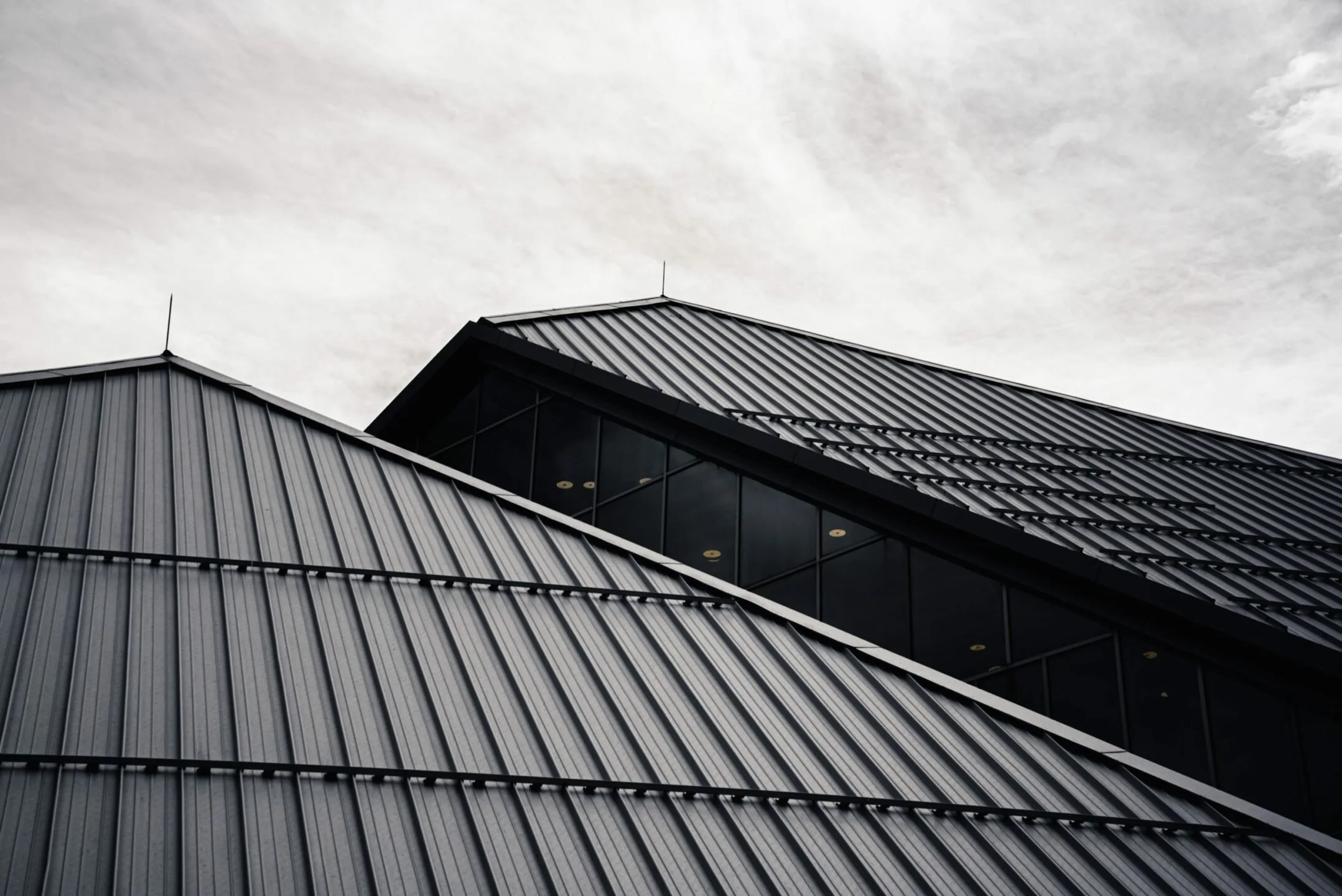 Close-up of a modern building with gray metal sloped roofs and large glass windows against a cloudy sky.